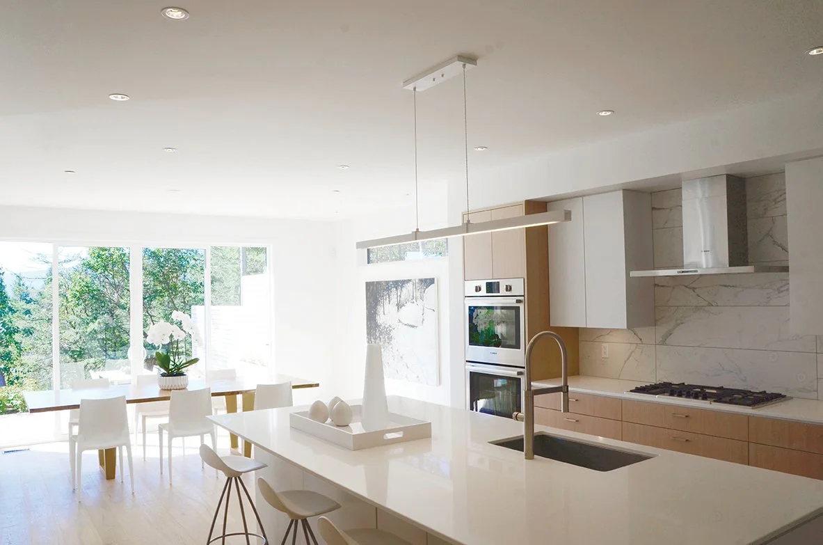Modern kitchen with white cabinets, a large island with a sink, and a dining area with white chairs and a wooden table with a flower arrangement. Large windows let in natural light and show greenery outside.