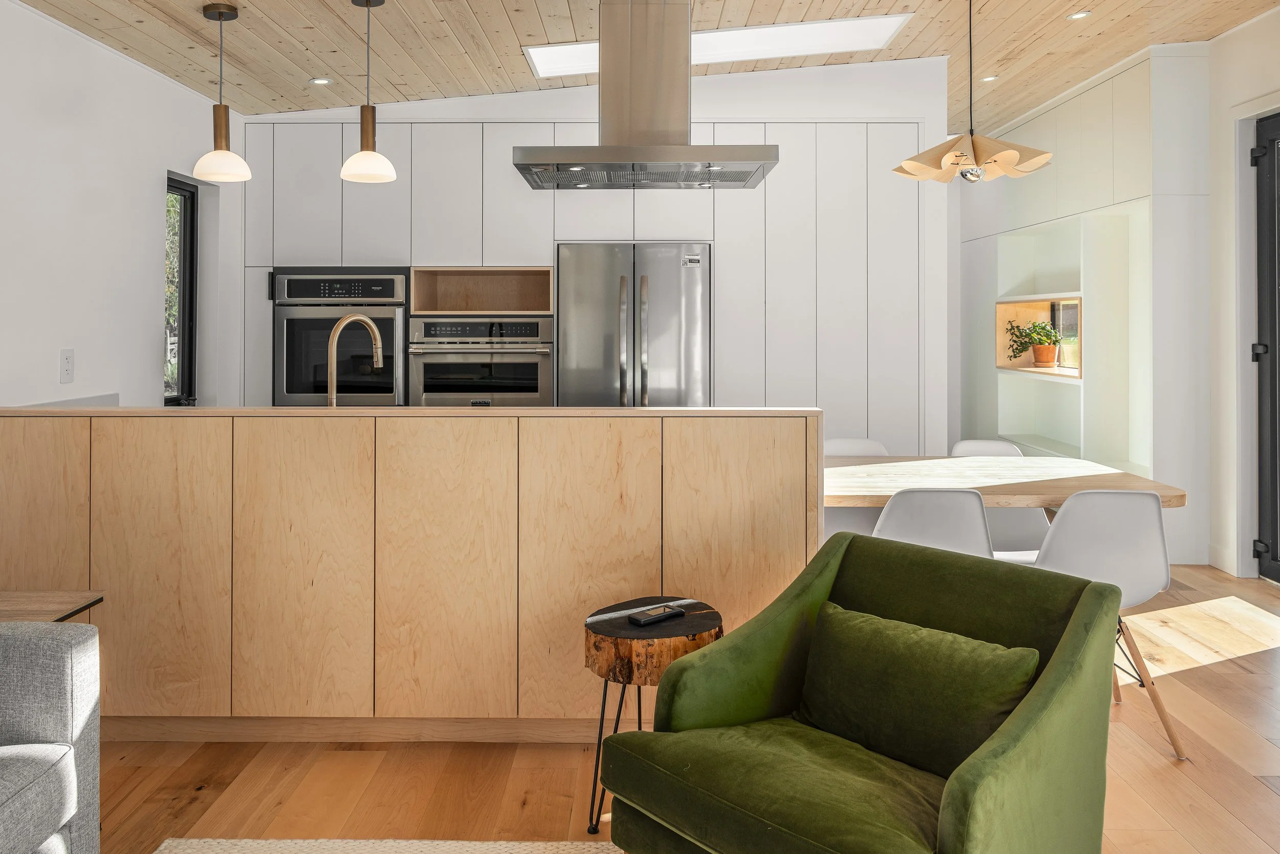 Modern open-concept kitchen and dining area with white and wood cabinetry, stainless steel appliances, a green velvet armchair, and a wooden dining table with white chairs.