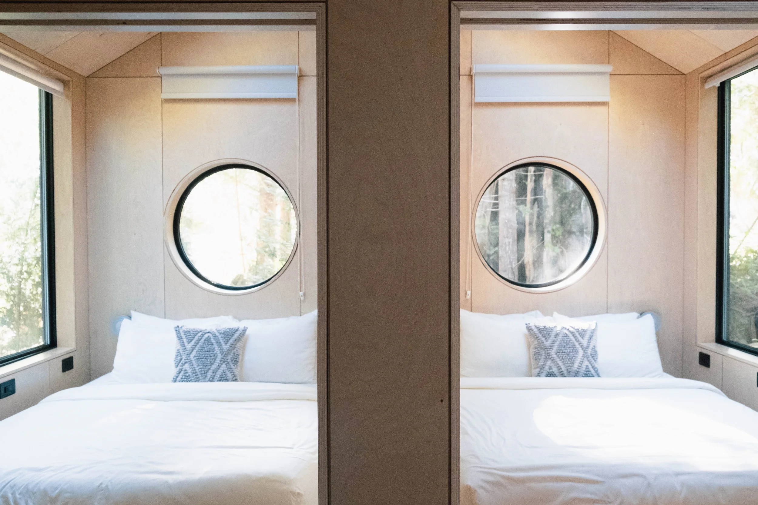 A mirrored image showing a bedroom with a bed, white bedding, decorative pillows, wooden walls, round windows, and large corner windows with views of trees outside.