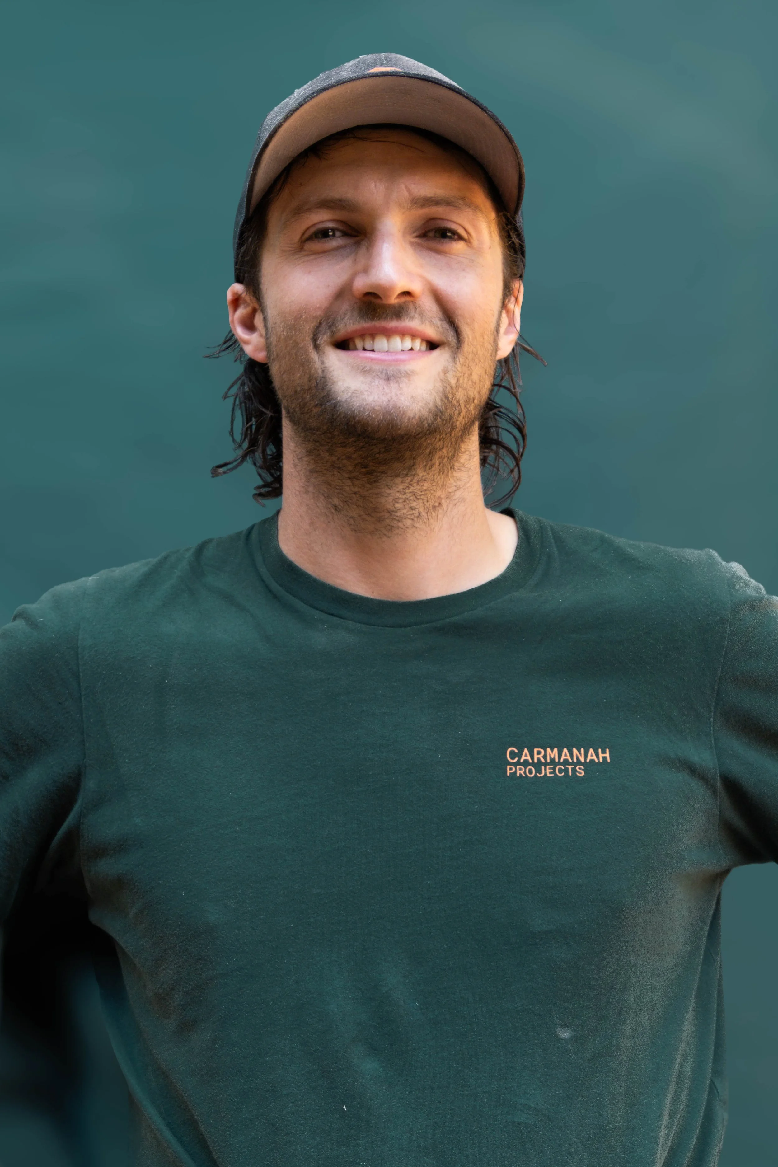 A young man wearing a dark cap and a dark green t-shirt with orange text, smiling confidently with arms crossed against a plain, dark background.
