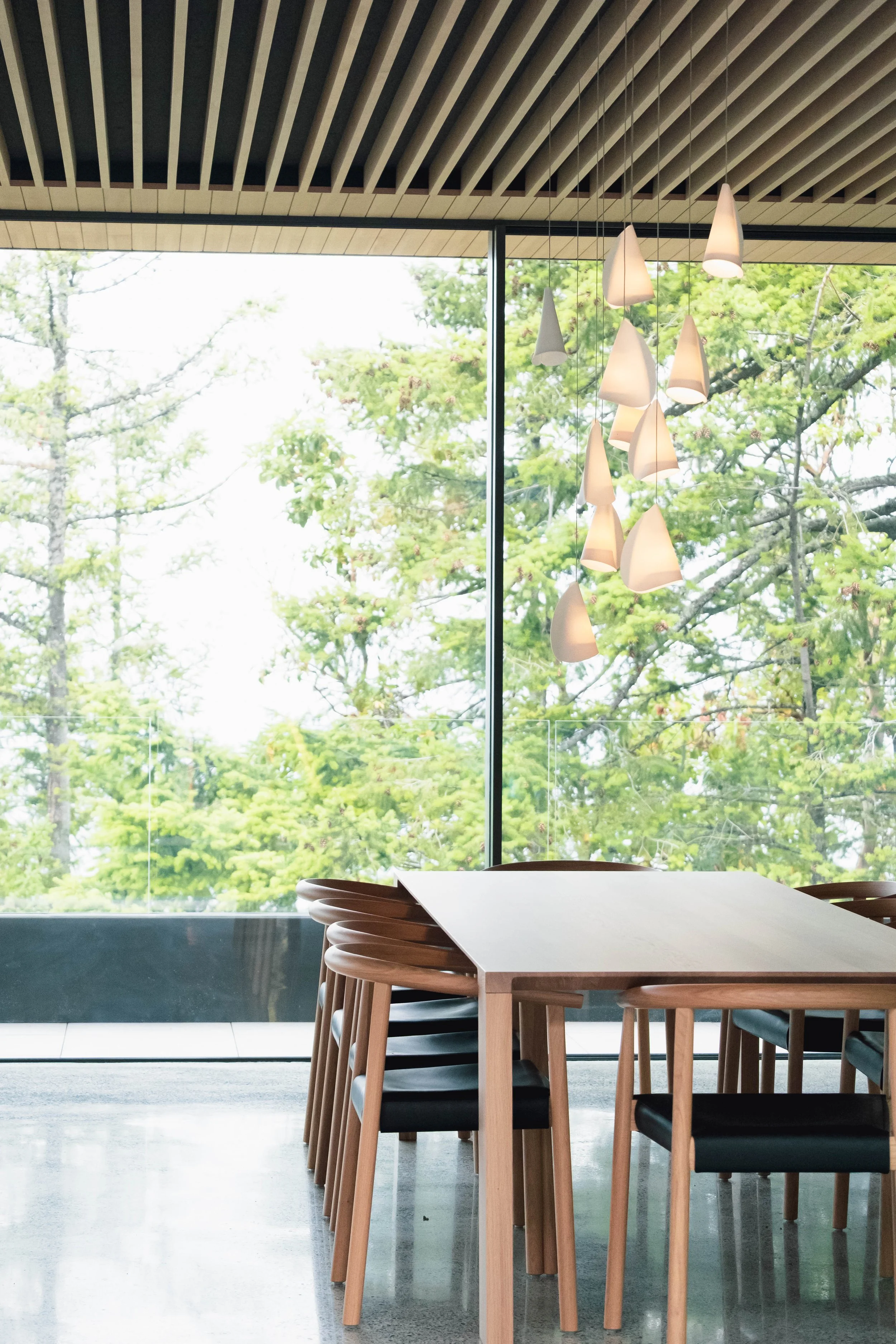 Modern dining area with wooden tables and black chairs, large floor-to-ceiling window showing green trees outside, and hanging pendant lights. 