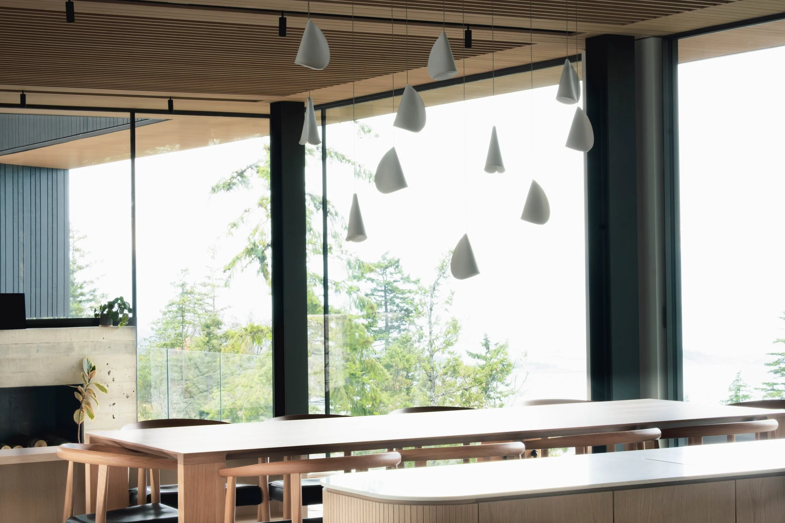 Modern interior with wooden ceiling, pendant lighting, large windows showing green trees, and a dining area with a wooden table and chairs.