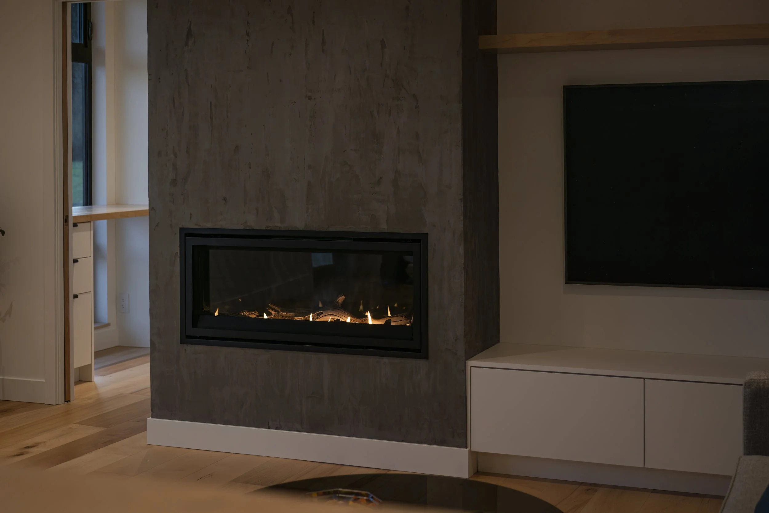 Modern living room with a cozy electric fireplace built into a textured gray wall, white entertainment console below, and a large flat-screen TV on the right. Part of a room with wooden floors and a small glimpse of an adjoining space.