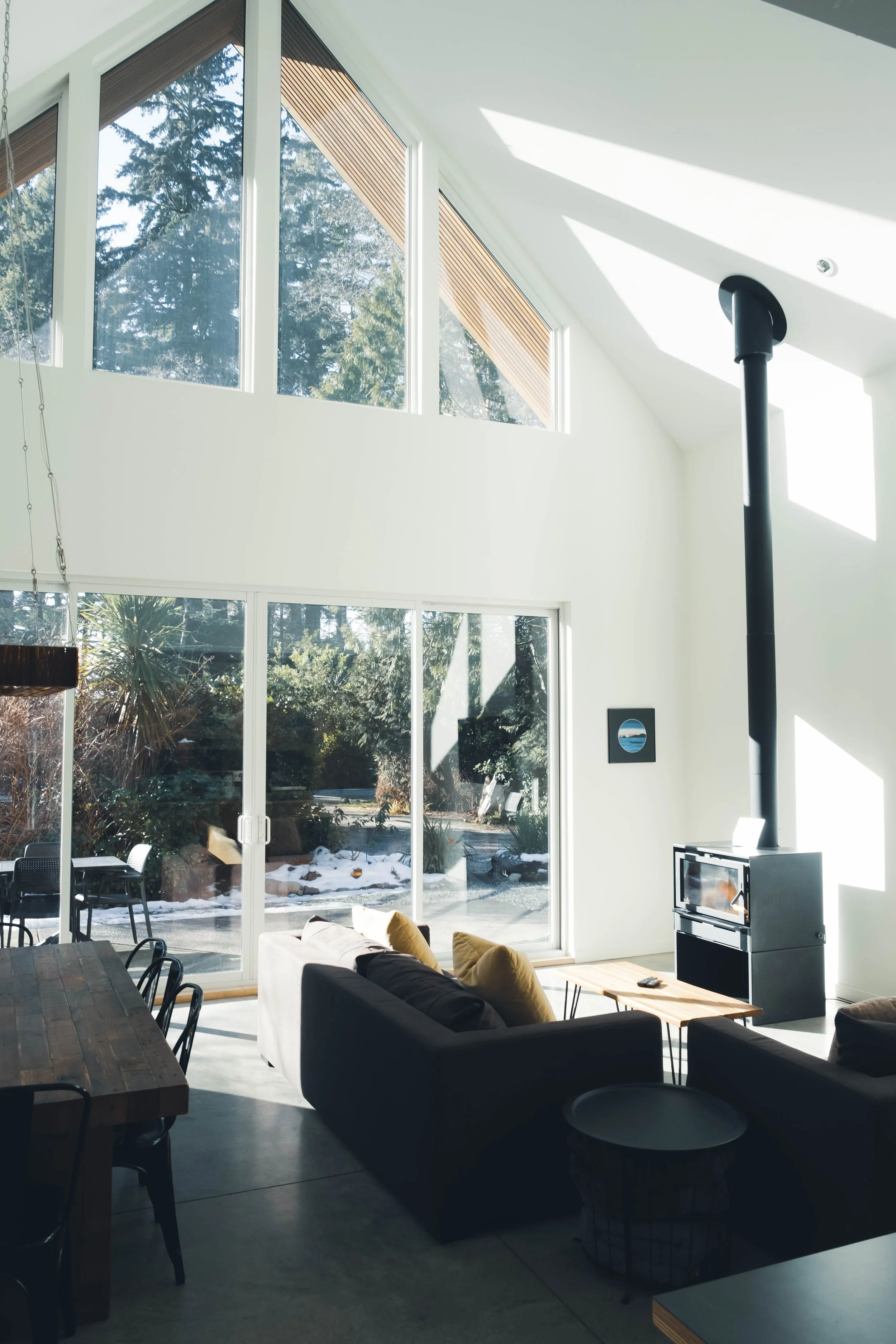 Living room with large floor-to-ceiling windows showing outdoor landscape, including snow and trees. Interior features a dark sofa with yellow cushions, a wooden coffee table, and a black wood stove with a chimney. There is a dining table with black 