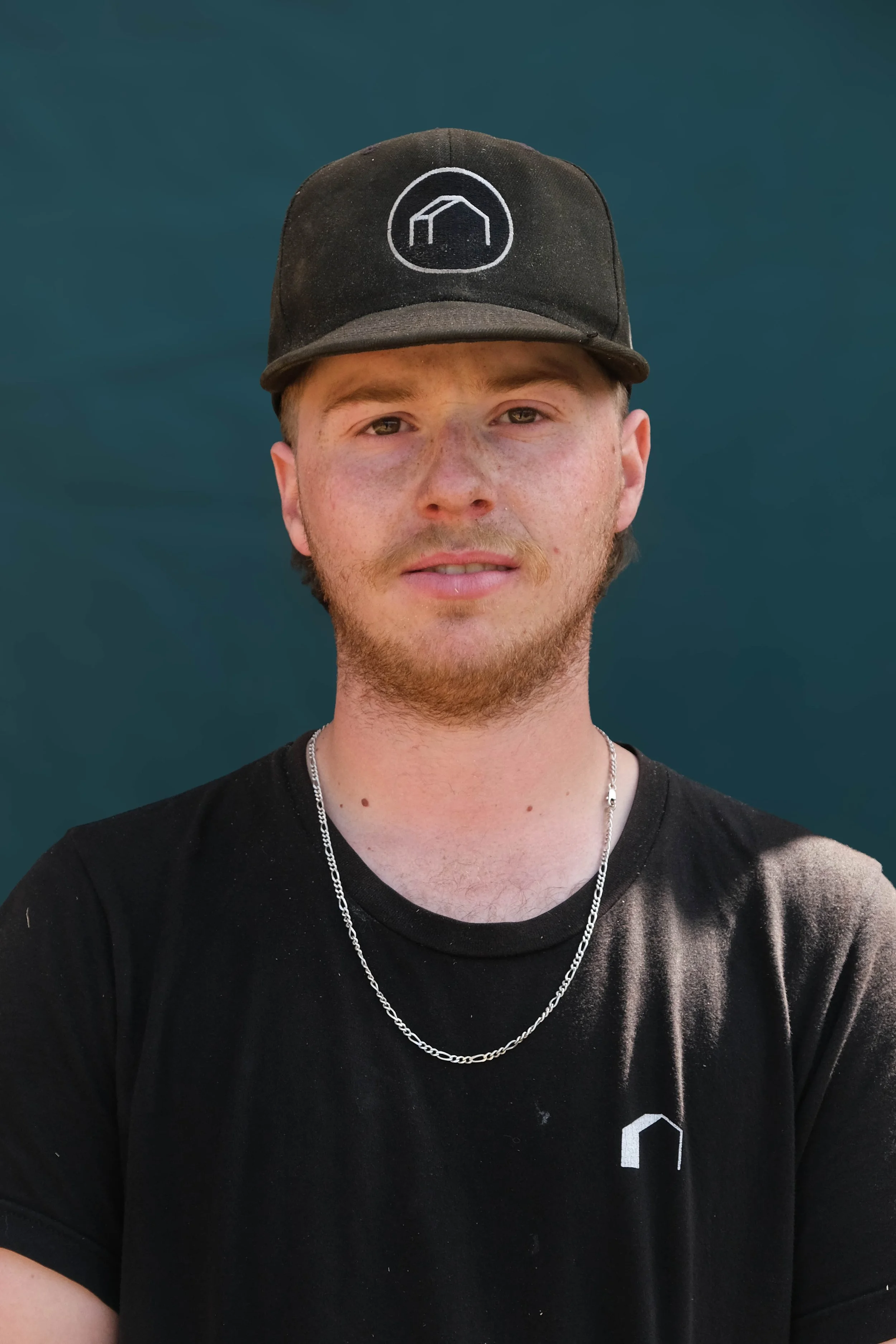 A young man with light skin, short reddish hair, and a beard, wearing a black baseball cap, a silver chain necklace, and a black T-shirt with a small white logo, standing against a dark blue background.