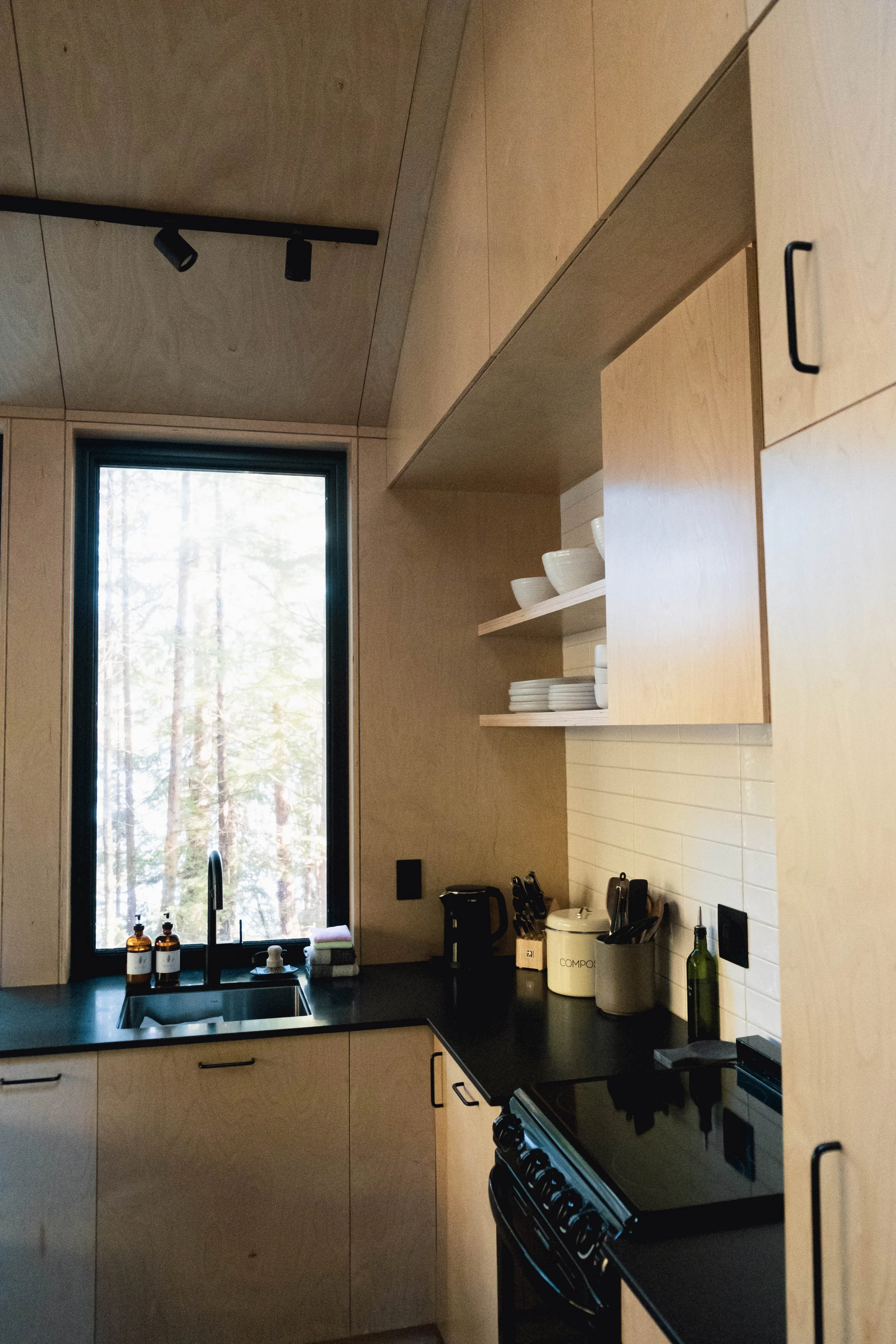 Kitchen with light wood cabinets, black countertop, a window overlooking trees, and various kitchen appliances and utensils.
