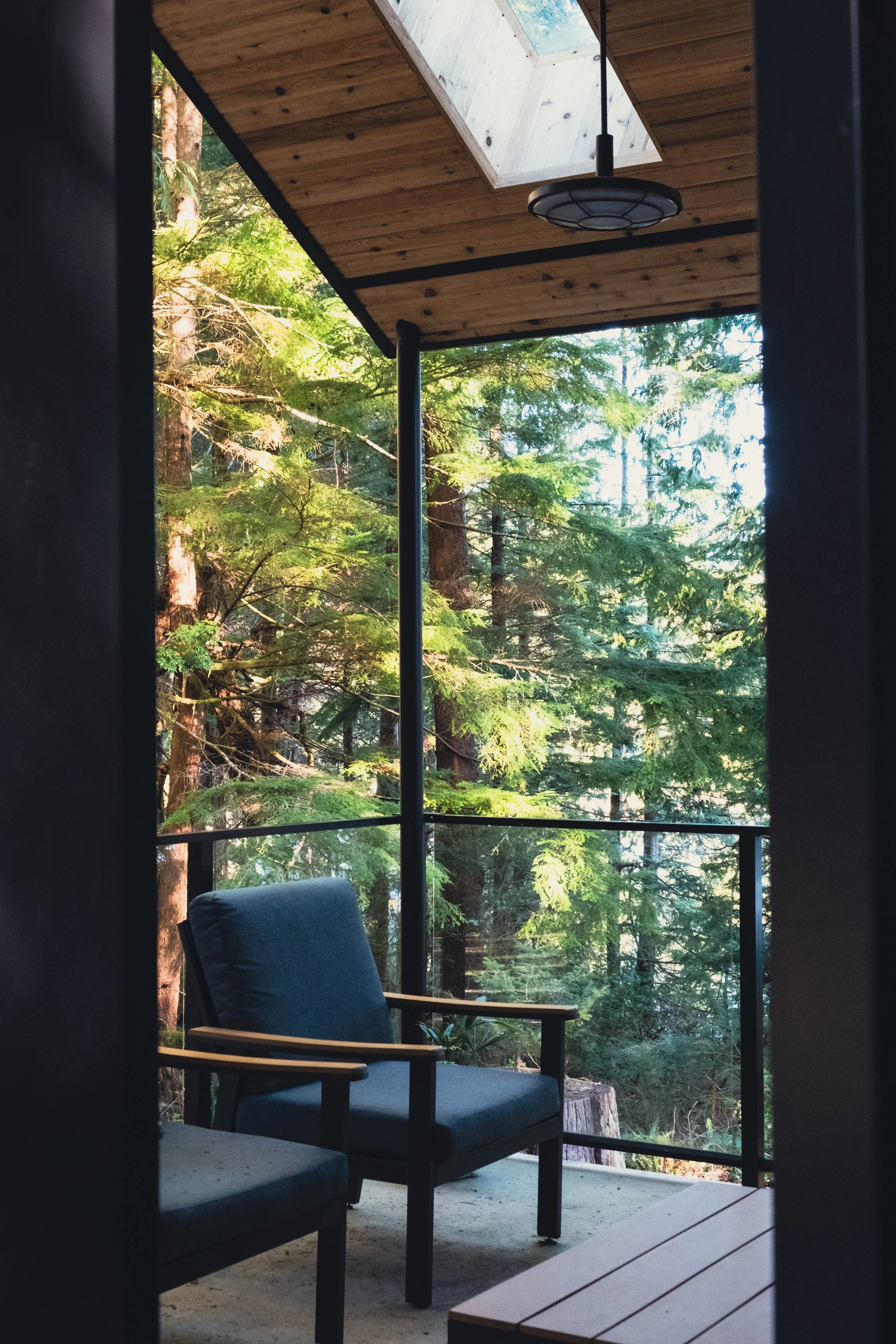 A porch with a black metal railing, two blue cushioned chairs with wooden armrests, a small wooden table, and a wooden ceiling with a skylight and a hanging light fixture, overlooking a forest of tall trees.