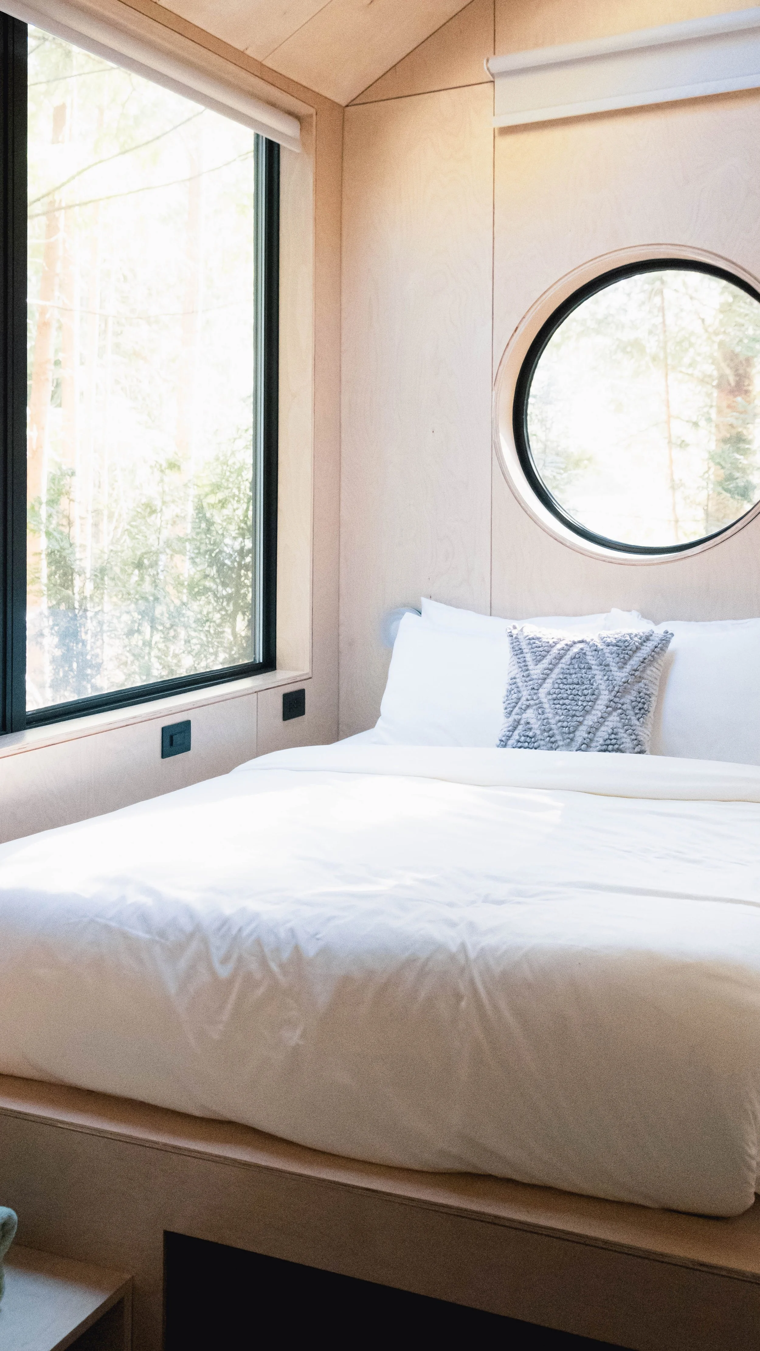 A cozy bedroom with a large white bed, decorative pillow, window with a view of trees, and round window on the wooden wall.