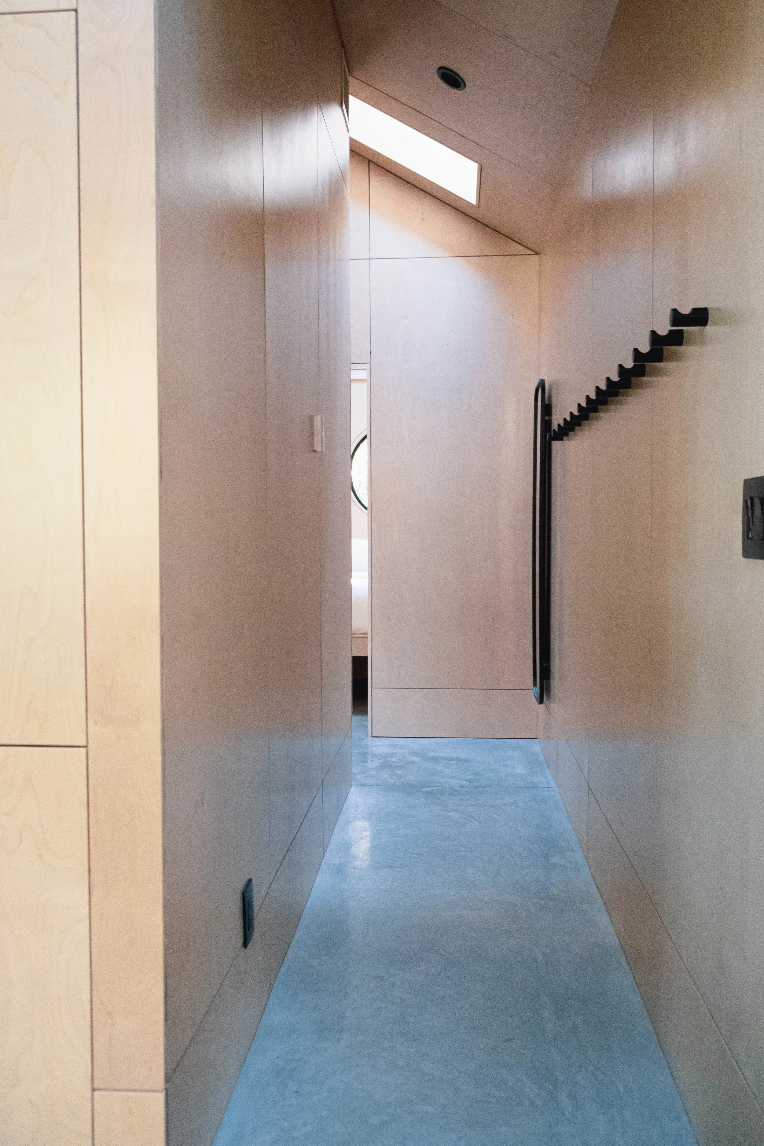 A narrow hallway with light wood walls and a gray concrete floor, leading to a room with a bed and circular window visible through an open door.