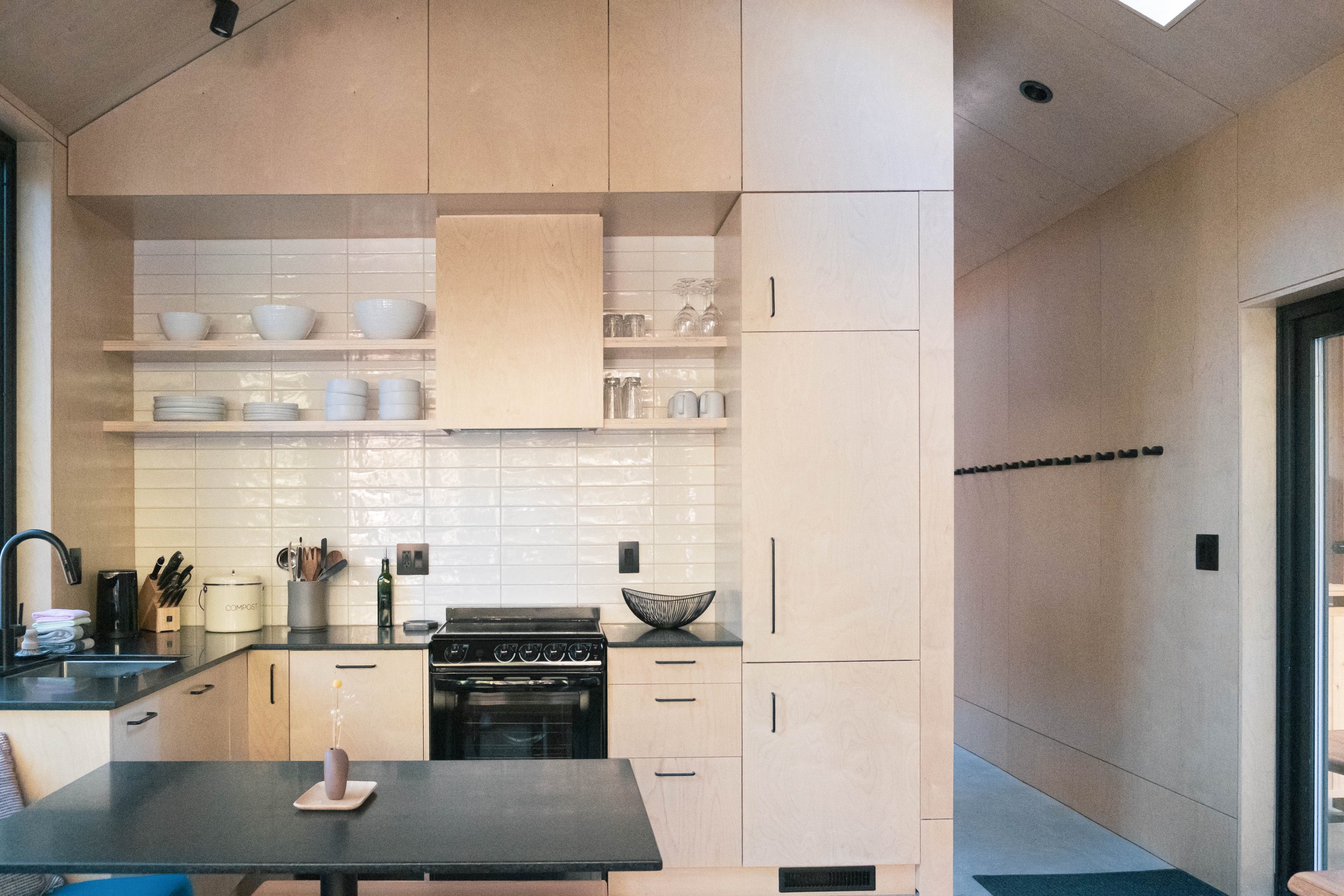 Modern kitchen with light wood cabinets, black countertops, white backsplash tiles, black stove, and open shelving with white dishes and glasses.