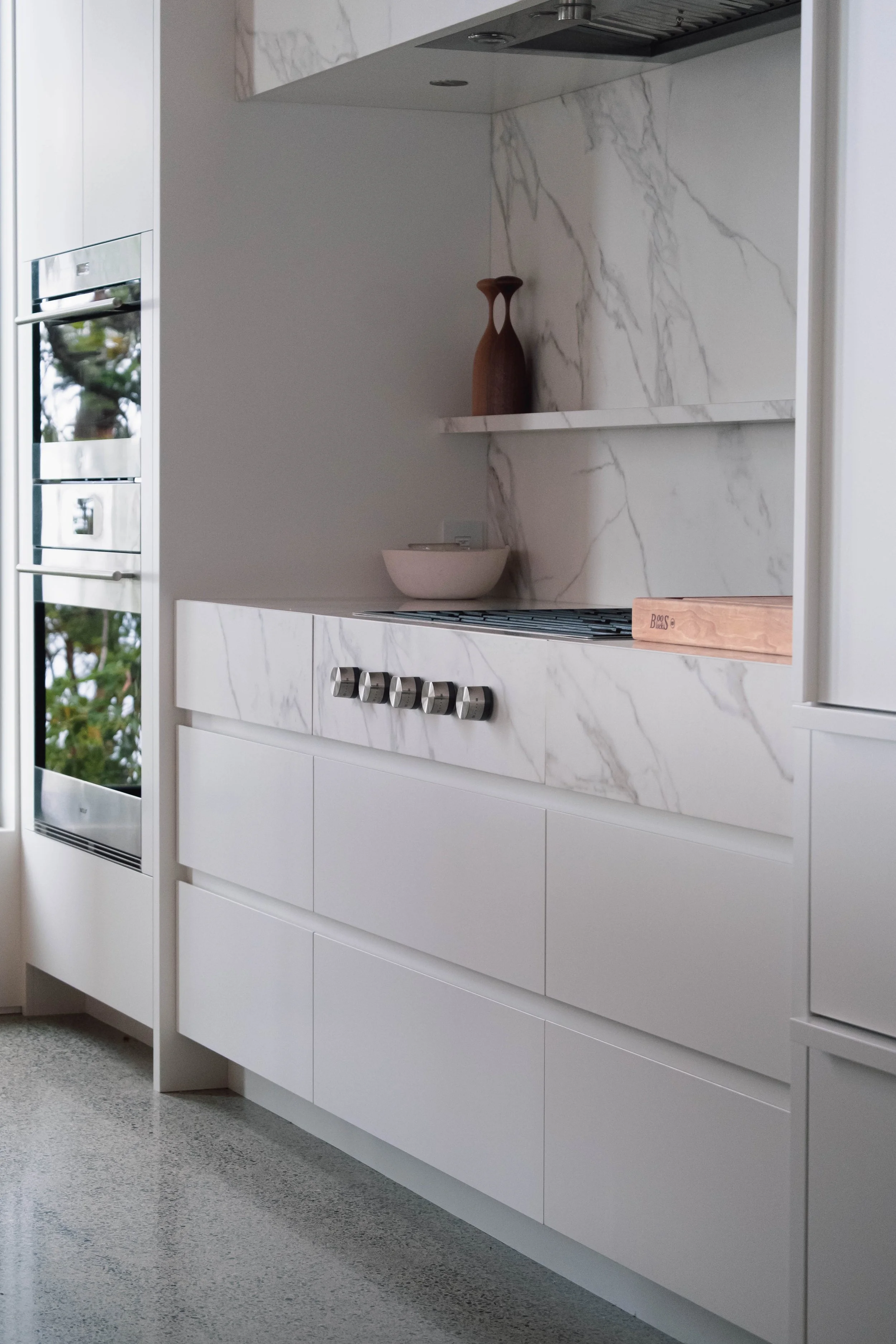 Modern white kitchen with marble countertops and backsplash, built-in oven, and minimal decor including a wooden vase, bowl, and a wooden tray.