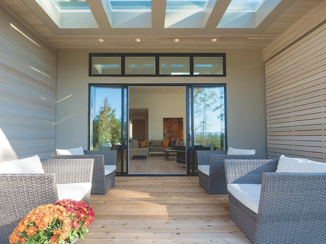 A modern outdoor patio area with wicker chairs and a potted plant with flowers, leading into a living room with a sofa and artwork through glass sliding doors, under a skylight ceiling.