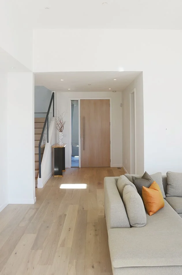 Modern living room with beige sofa and colorful pillows, light wood flooring, white walls, staircase to the left, and a wooden front door in the center.