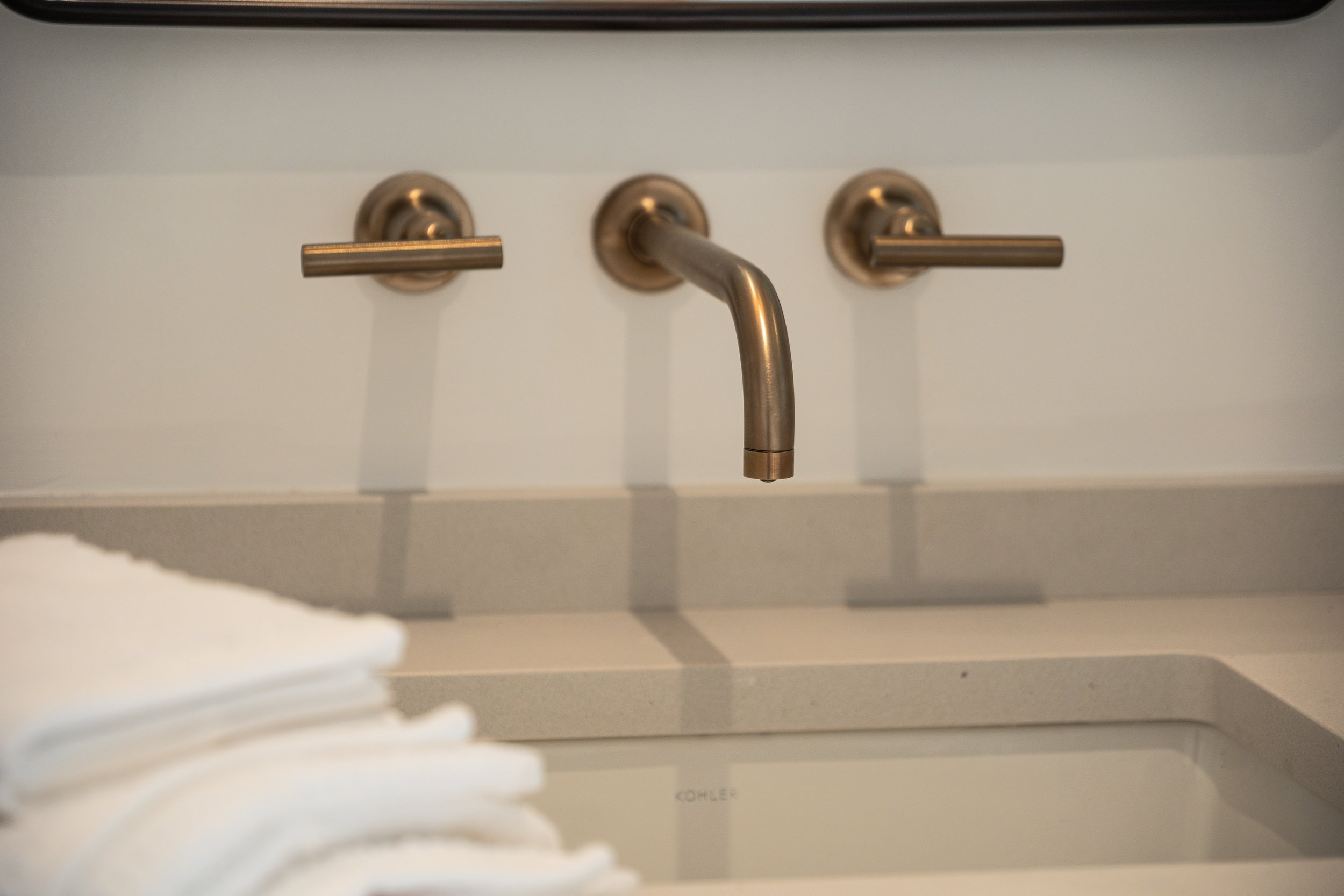 Close-up of a wall-mounted modern sink faucet with two levers, with a white towel in the foreground and part of a white sink visible at the bottom.