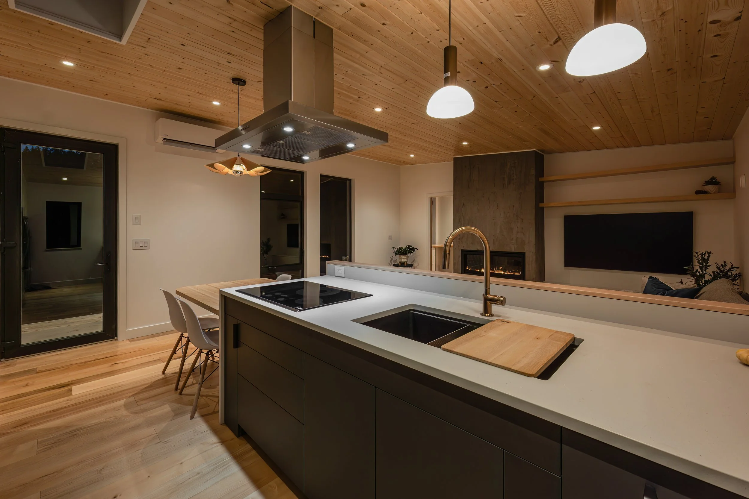 Modern kitchen with wooden ceiling, pendant and recessed lighting, black cabinetry, white countertop, dual sink, induction stove, and wooden cutting board, open to a living area with a fireplace and TV.