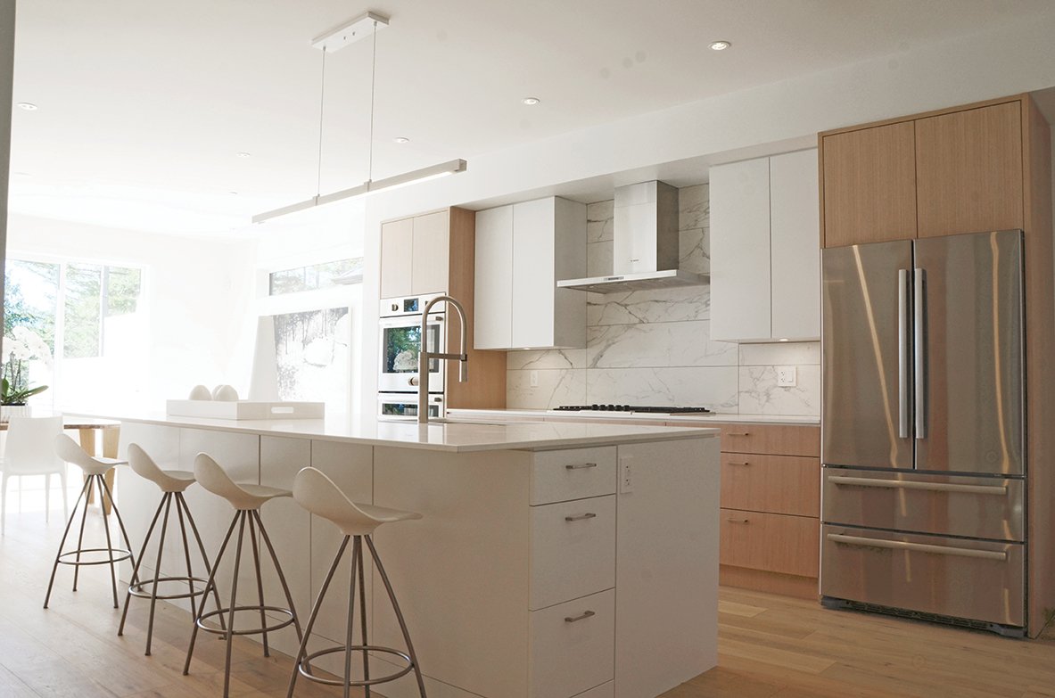 Modern kitchen with white cabinets, a marble backsplash, a stainless steel refrigerator, a kitchen island with white chairs, and a window letting in natural light.