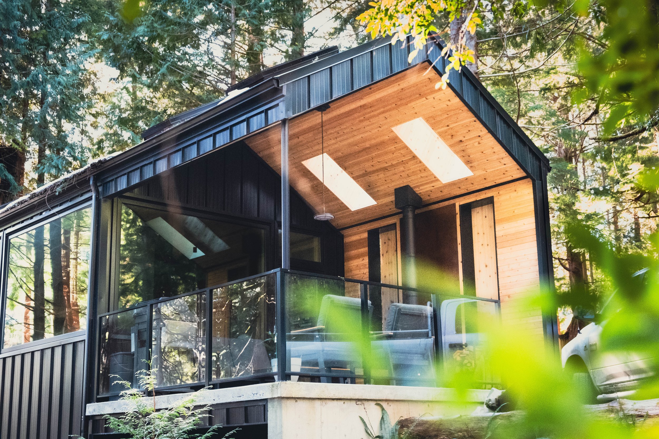Modern house with black exterior and wooden accents, surrounded by trees, featuring a glass balcony with furniture, in a forested setting.