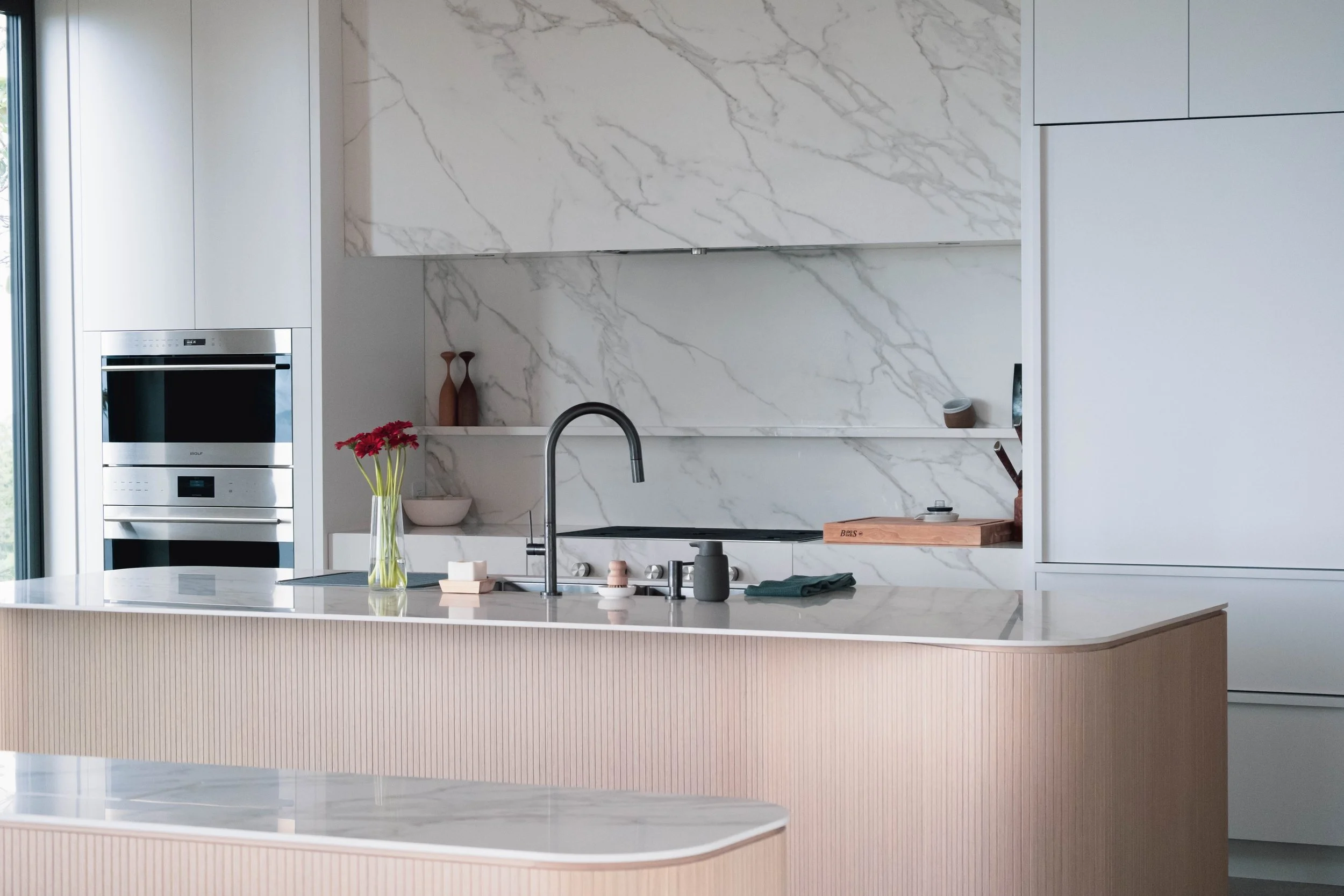 Modern kitchen with white marble backsplash, white cabinets, light wood island, and black faucet. Contains decorative vases, a vase with pink flowers, a bowl, and kitchen utensils.