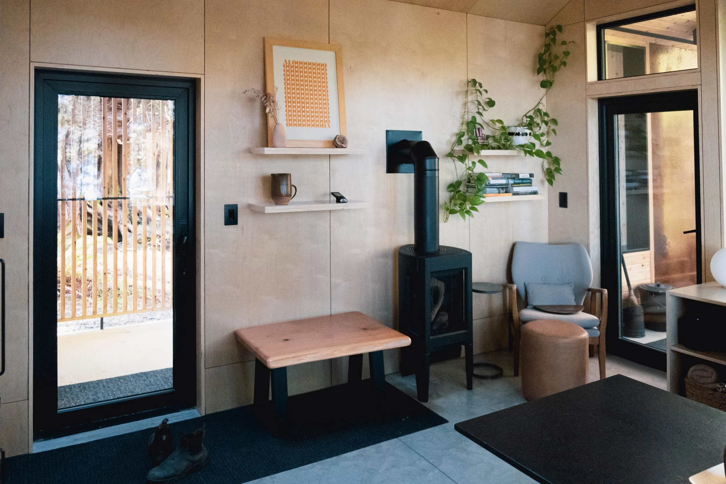 Cozy interior with a door leading outside, a wood stove, a comfortable armchair, a small stool, bookshelves with books, and plant decorations.