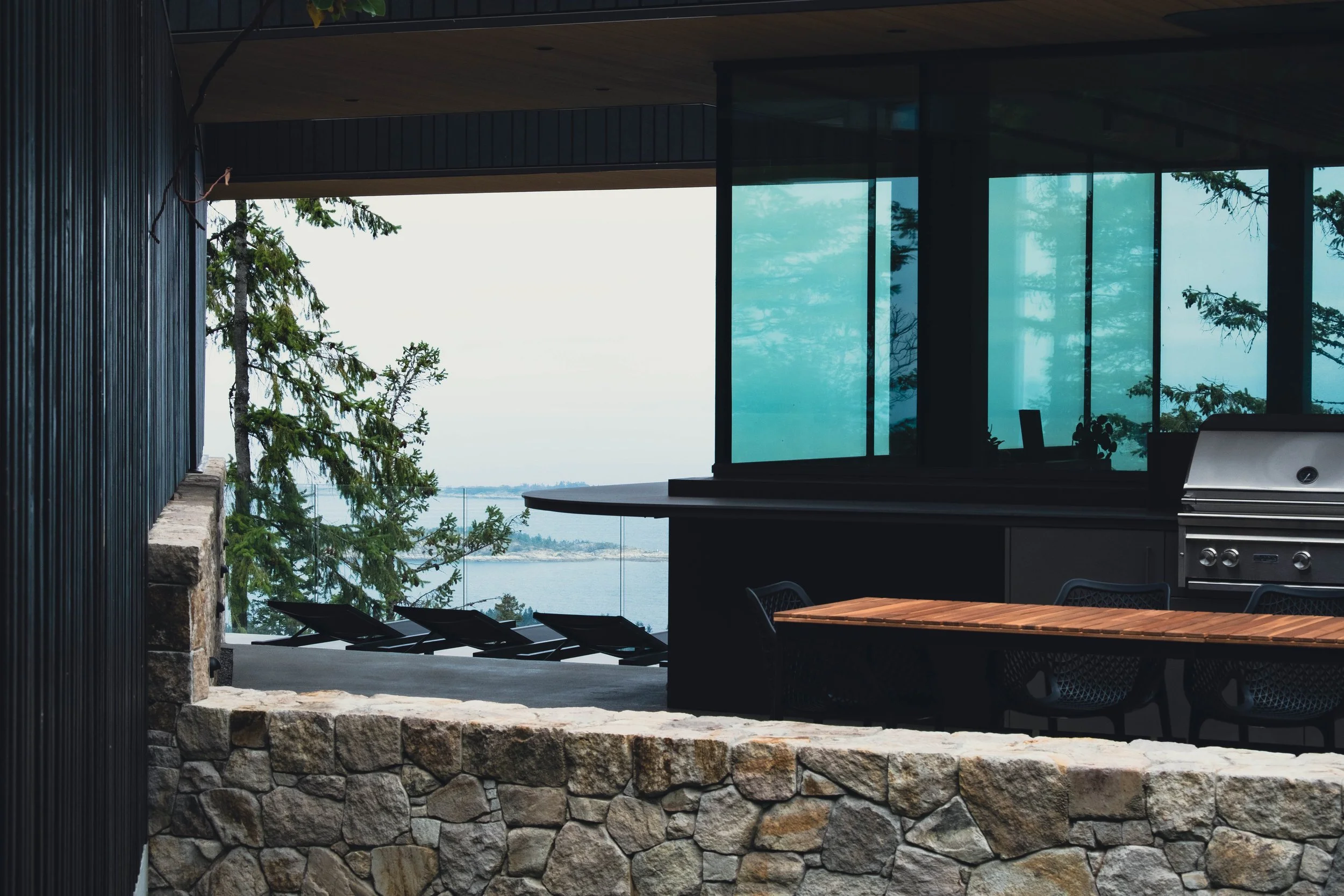 Modern outdoor patio area with stone wall, black lounge chairs, a wooden dining table, black chairs, and a barbecue grill, overlooking a body of water with trees.