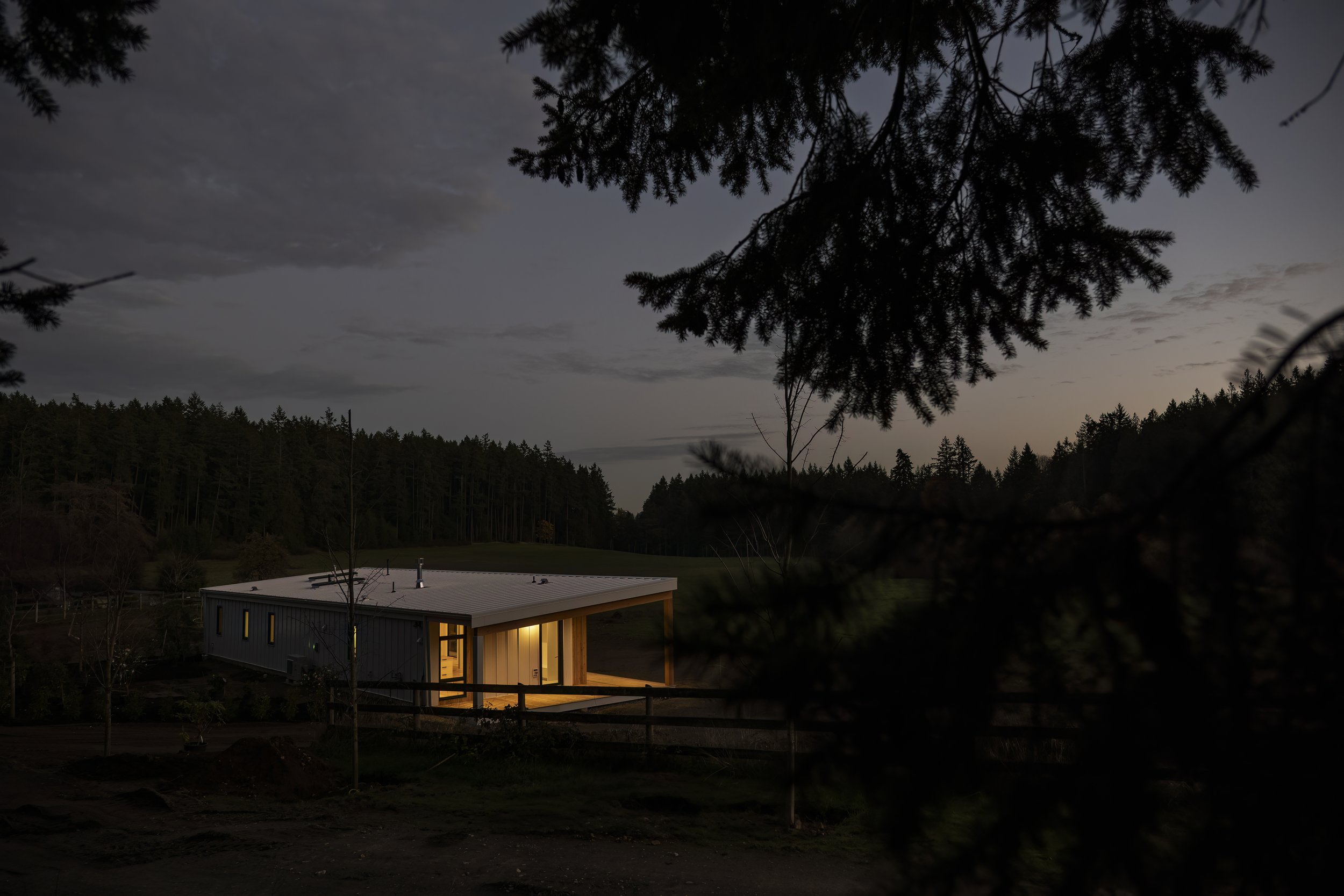 A modern house illuminated from within at dusk, surrounded by trees and a forest in the background.