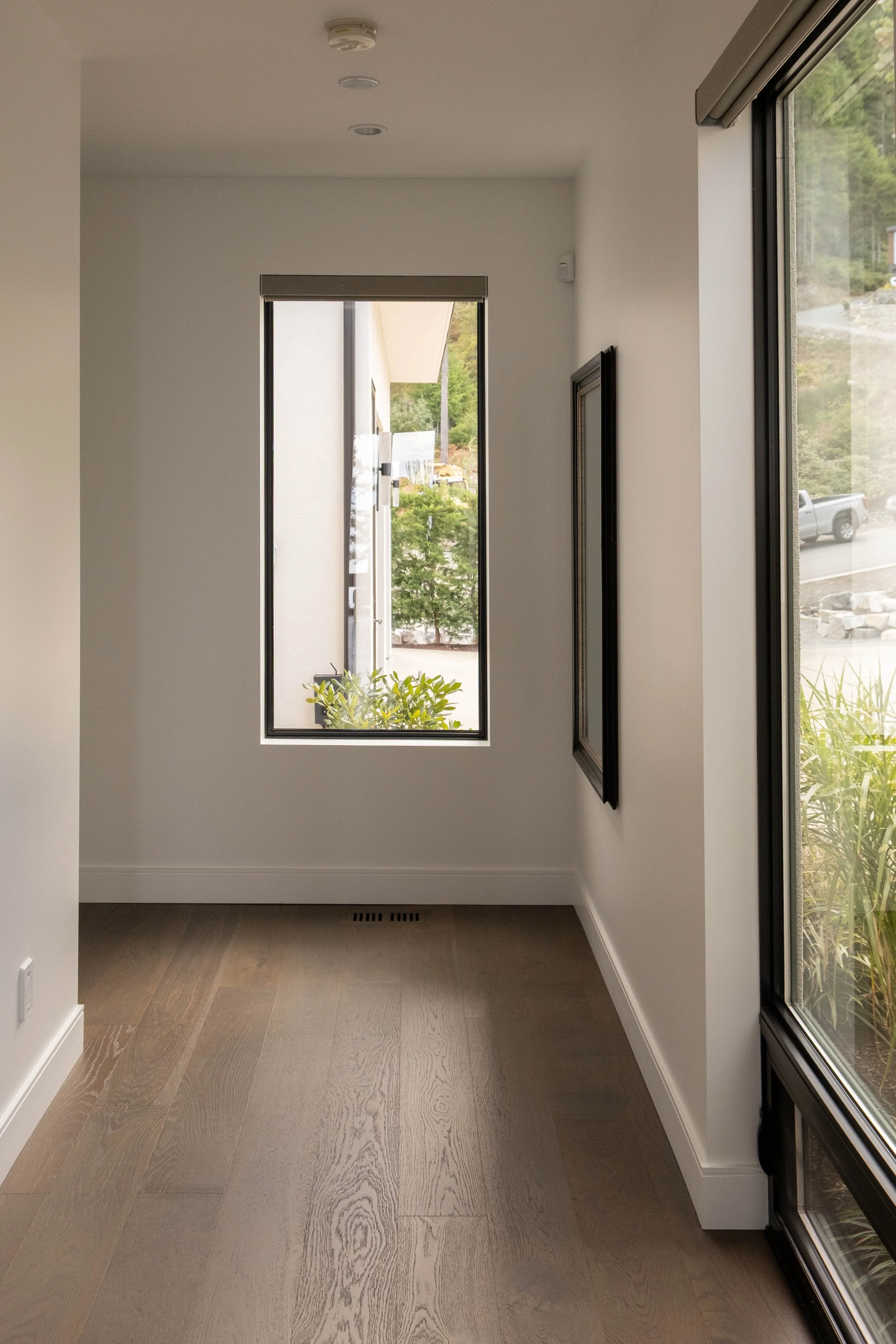 Empty room with wood flooring and large windows showing outdoor greenery and part of a street.