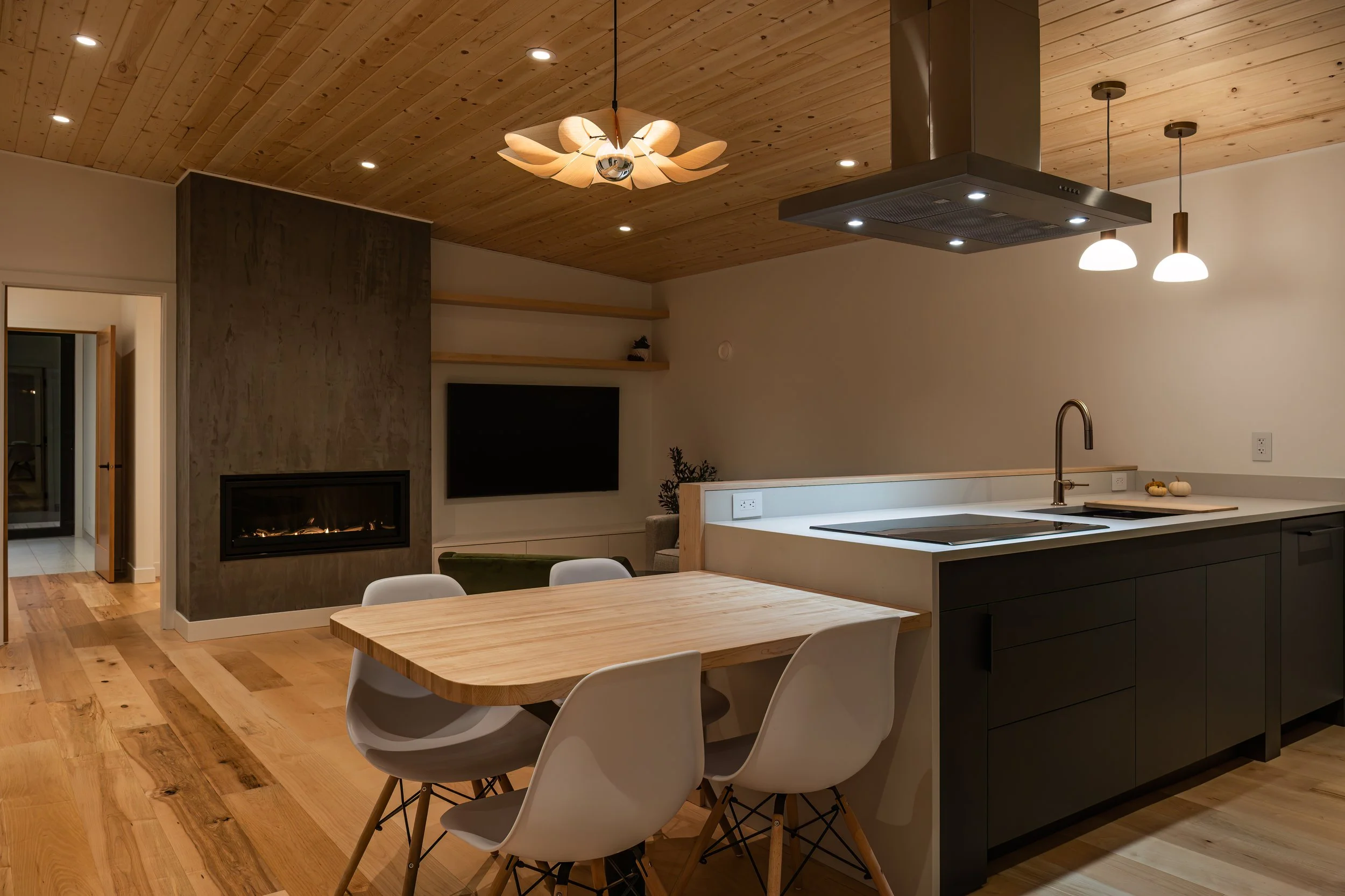 Modern kitchen with wooden ceiling, island with sink, black cabinets, dining table with white chairs, fireplace, and mounted television, illuminated by ceiling and pendant lights.