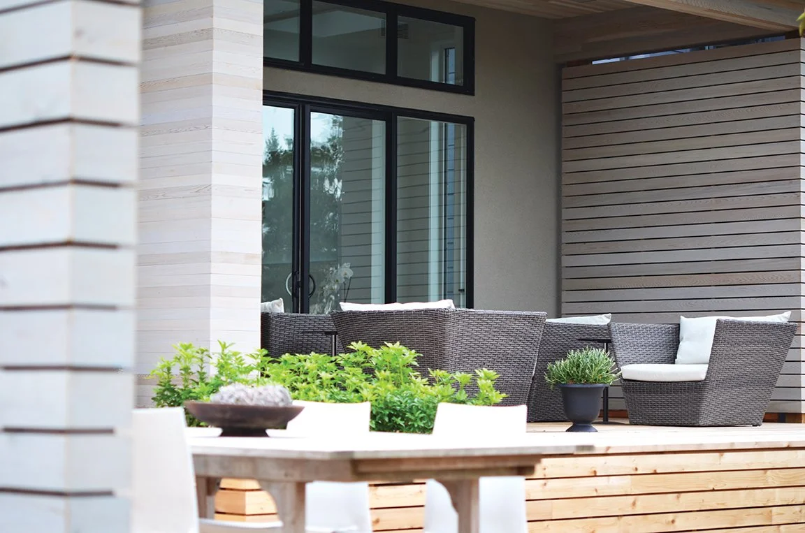 Outdoor patio area with wicker chairs, cushions, and potted plants, situated on a wooden deck outside a house with large glass sliding doors and modern siding.