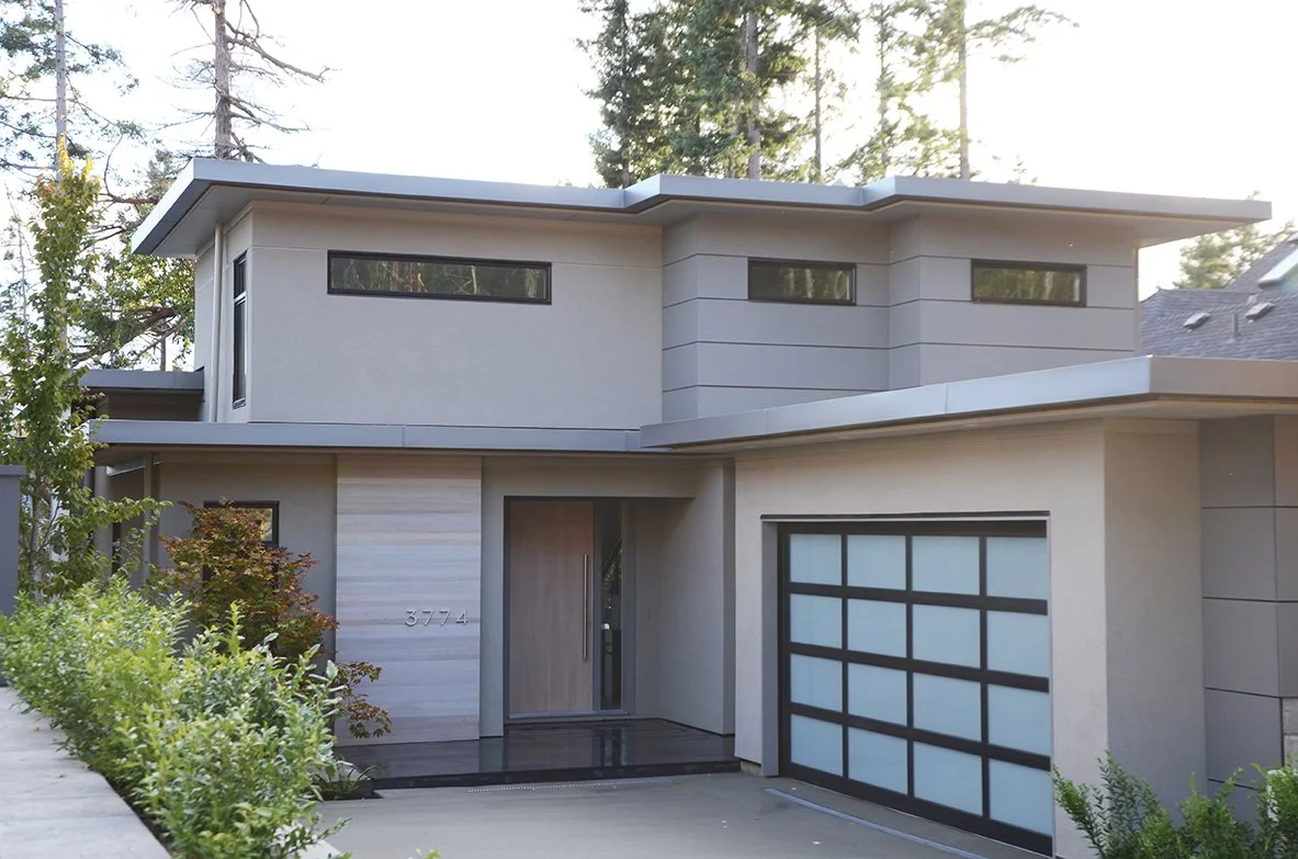 Modern two-story house with a flat roof, large windows, and a garage door, surrounded by greenery.