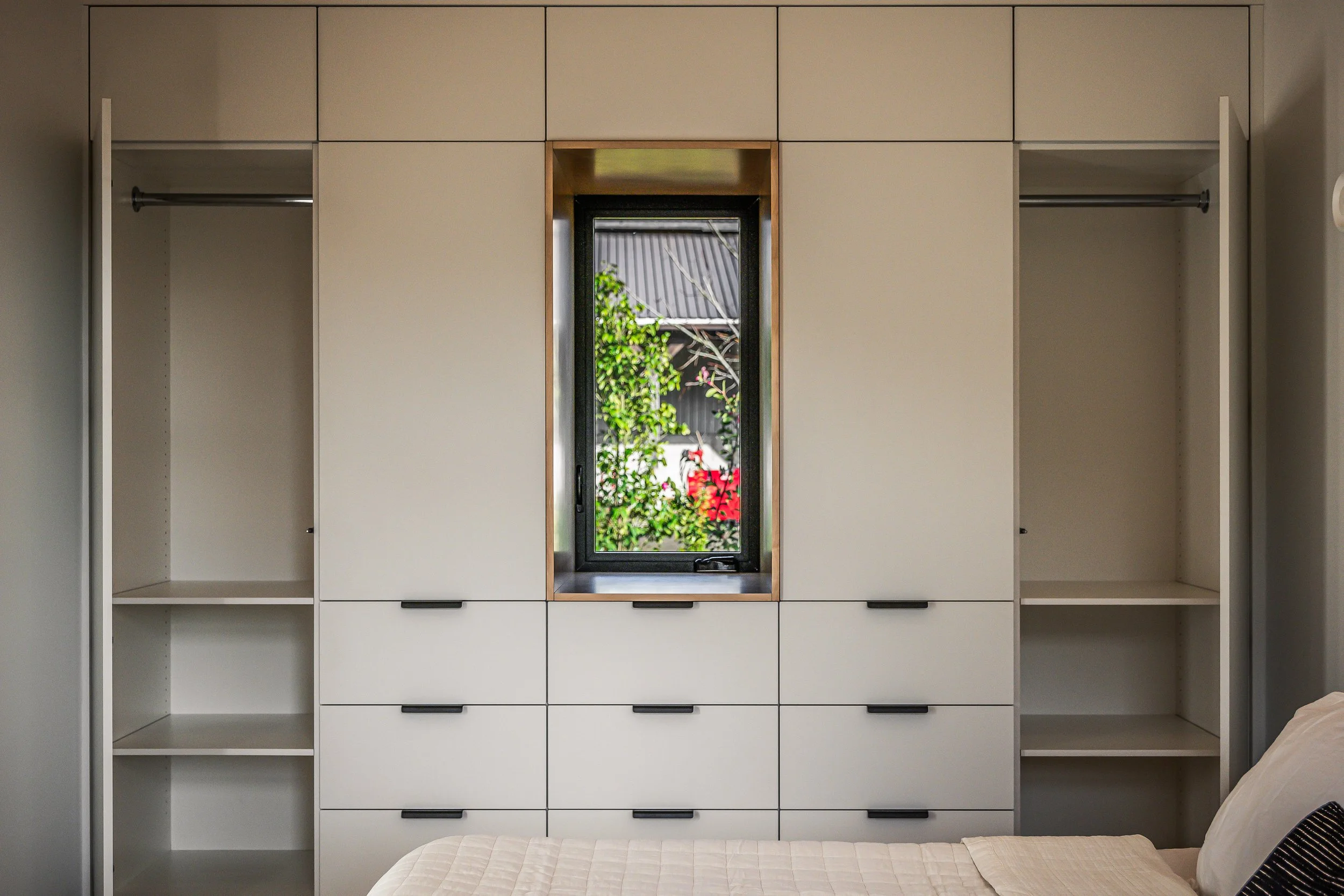 Built-in white wardrobe with open shelves and drawers, featuring a central narrow window with a view of trees and a roof outside.