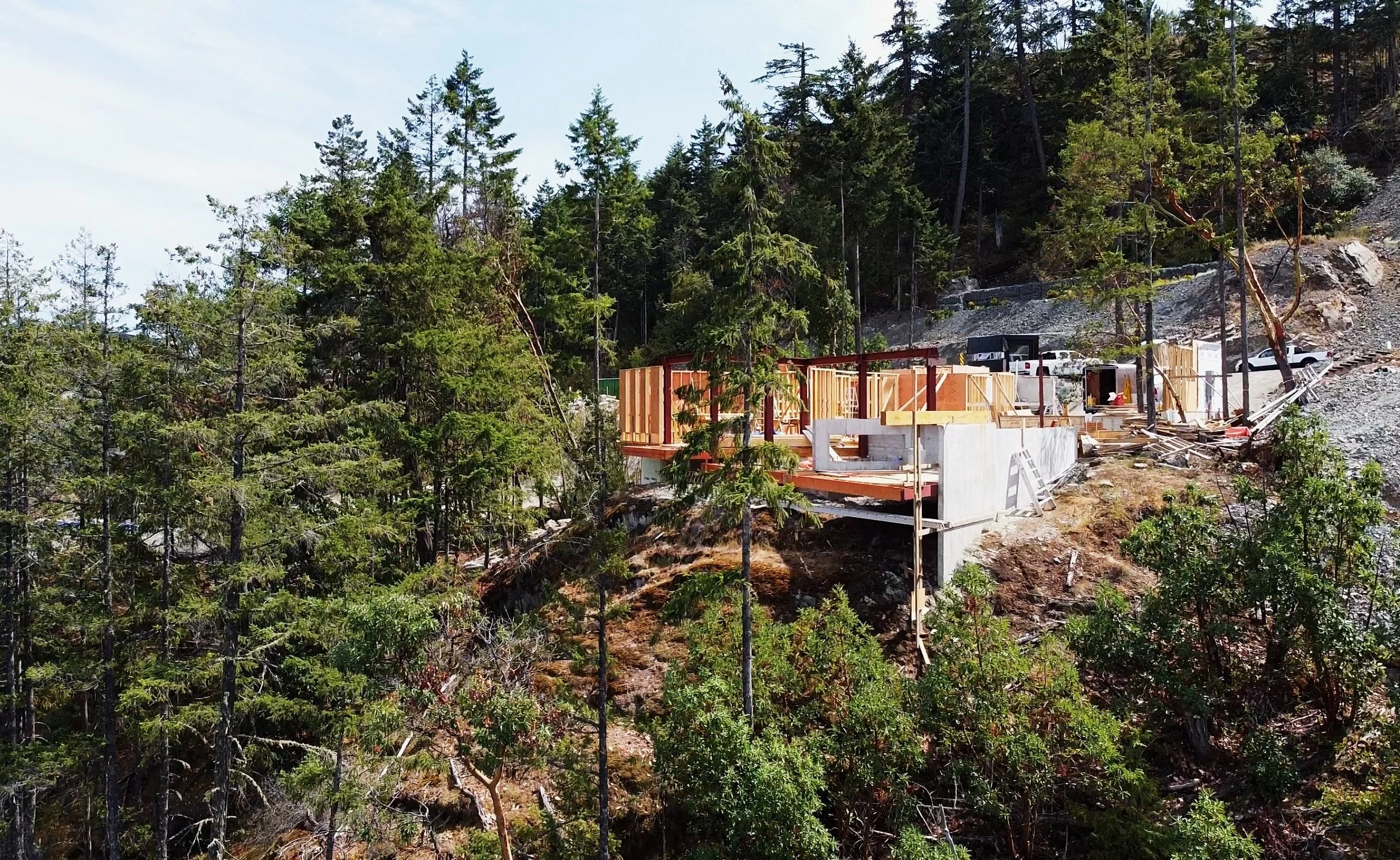 Construction site on a hillside surrounded by tall pine trees, with partially built structure and construction materials.