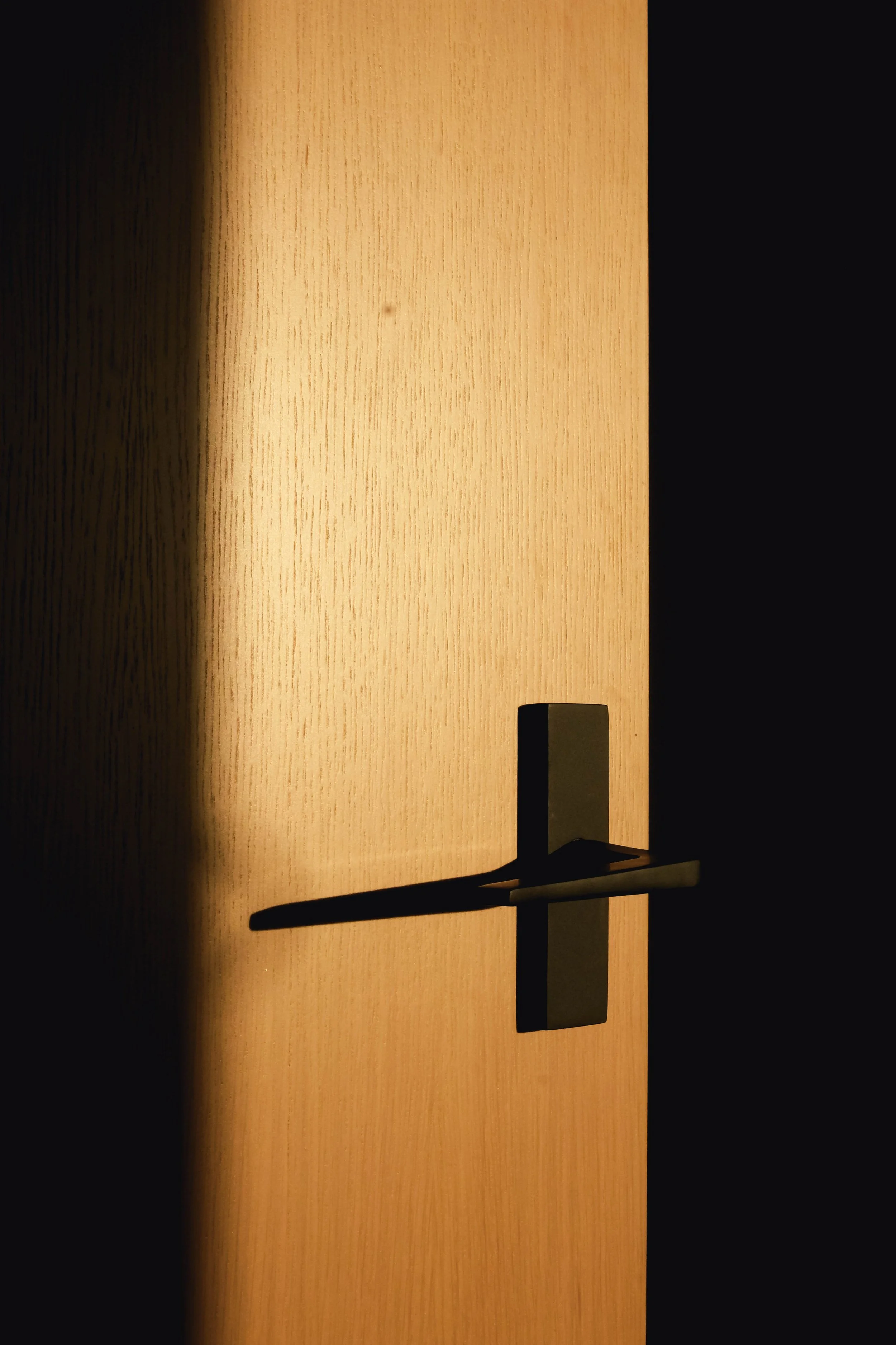 A close-up of a black door handle and lock mechanism on a wooden door with a warm light casting a shadow on the door surface.