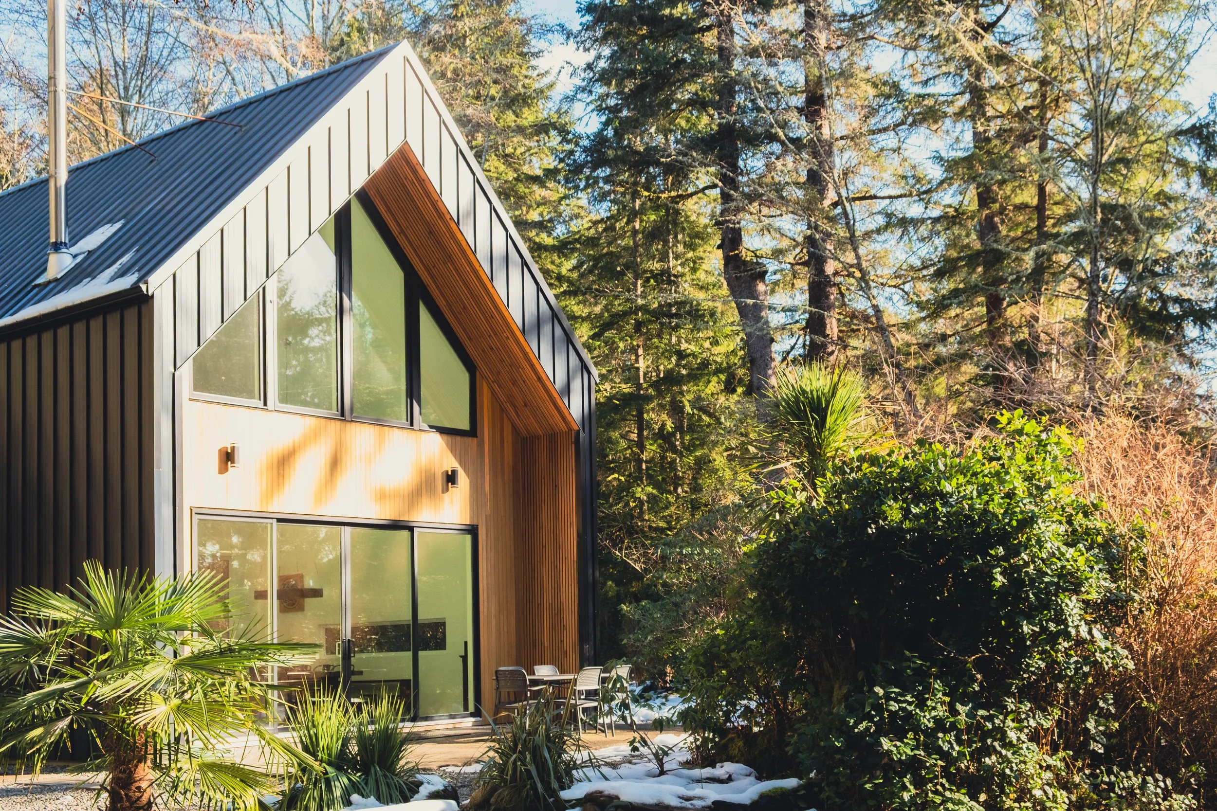 Contemporary house with a sharp, angular roof and large windows, surrounded by green plants and trees on a sunny day, with signs of snow on the ground.