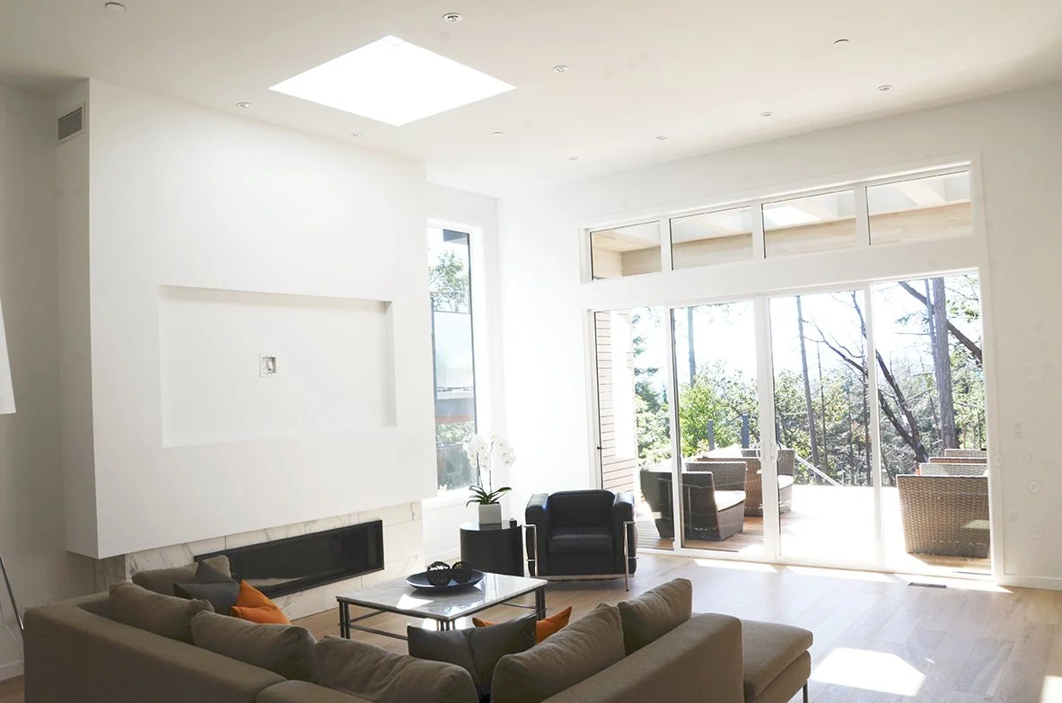 Modern living room with large glass sliding doors, outdoor patio with wicker chairs, beige sectional sofa, black armchair, coffee table, fireplace, and bright natural light.