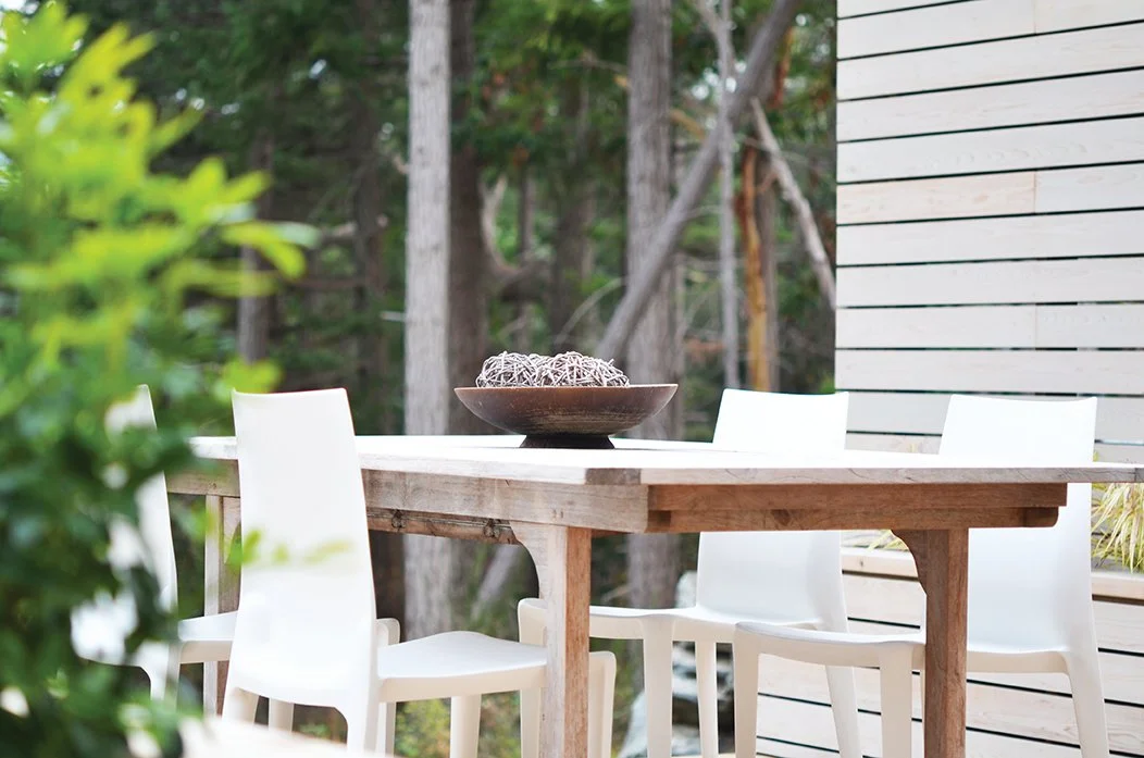 Outdoor dining table with six white chairs, a decorative bowl with woven balls, surrounded by trees and wooden siding.