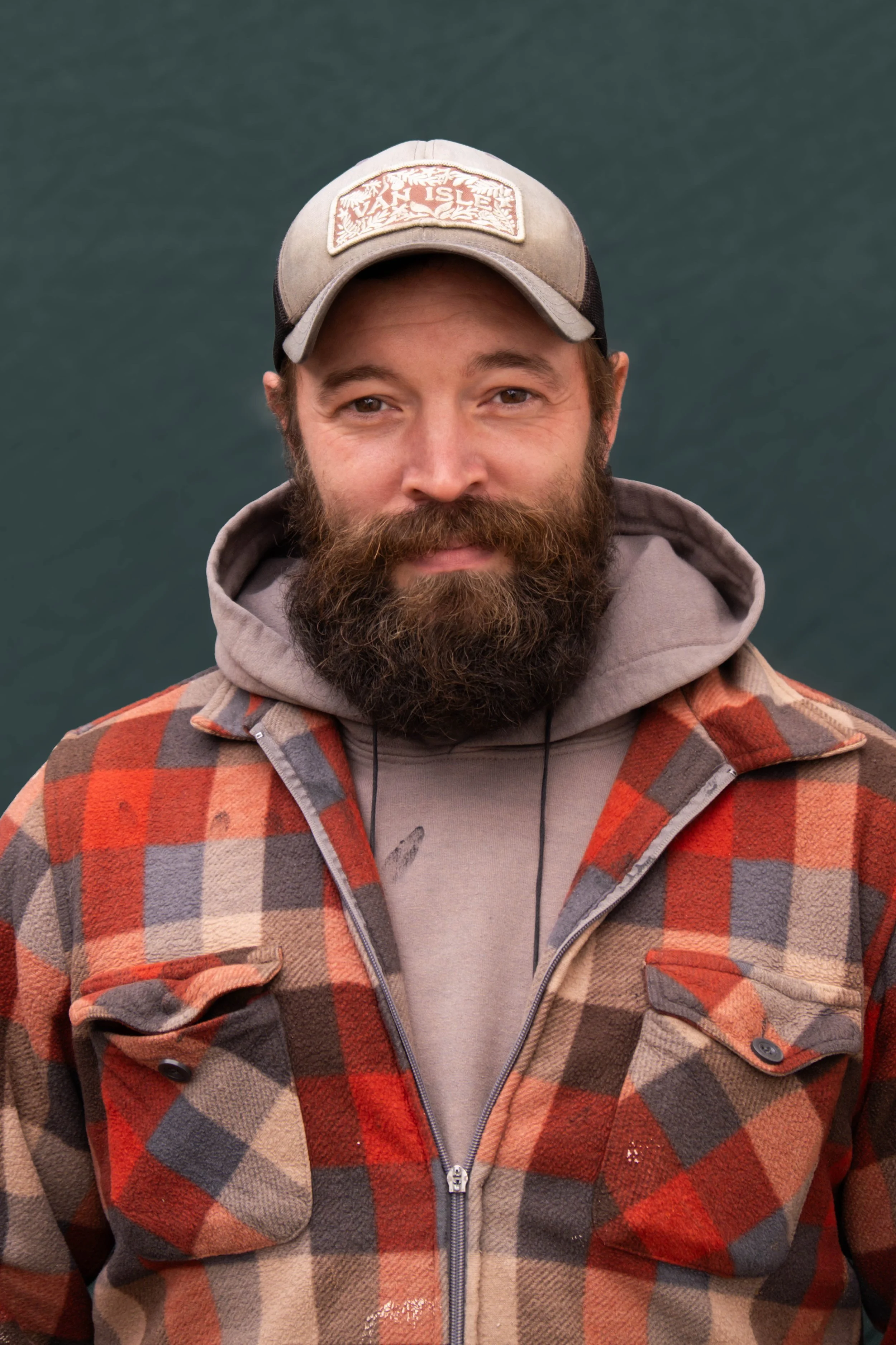 A man with a beard wearing a gray cap, beige sweatshirt, and a plaid jacket standing outdoors.
