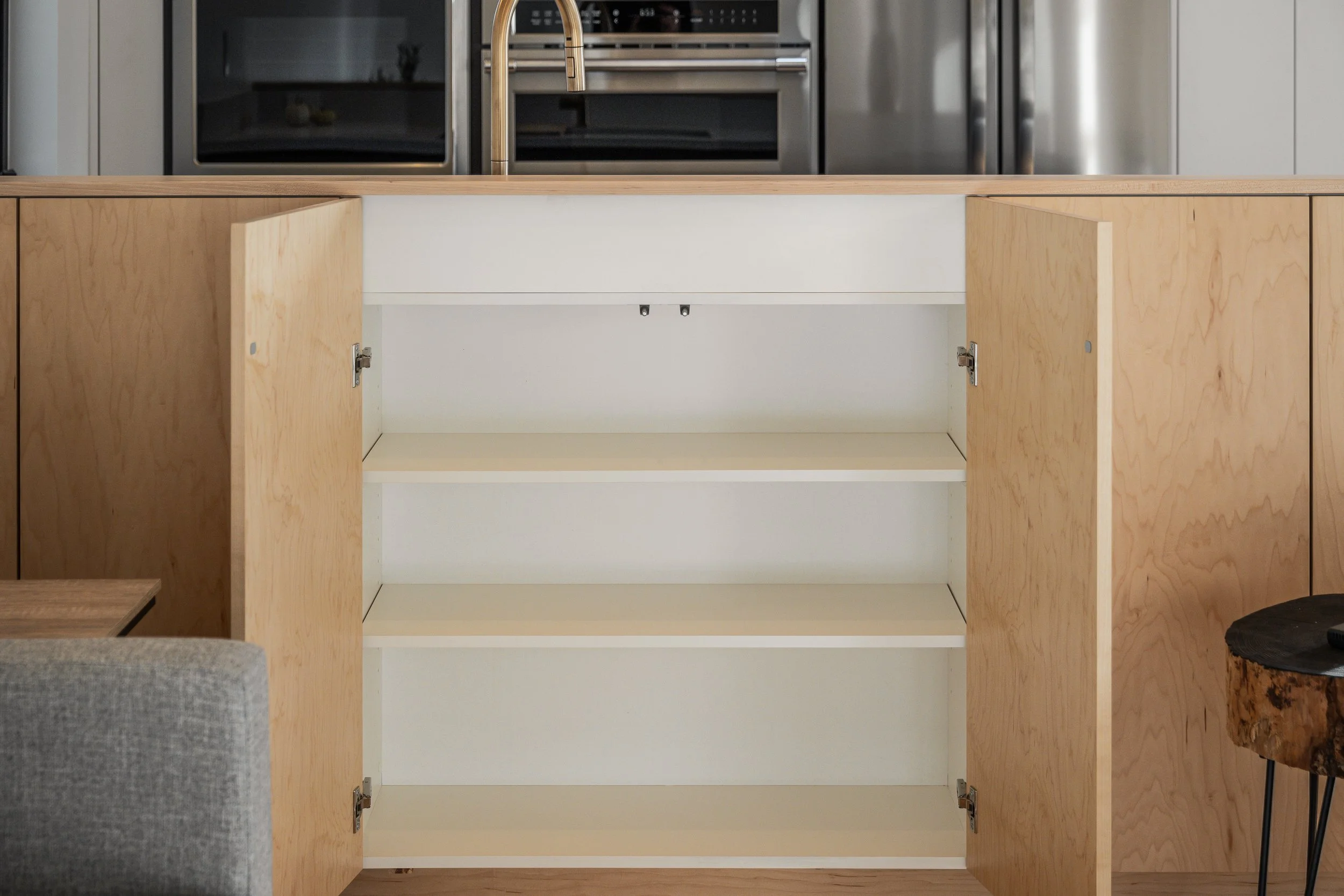 Open wooden kitchen cabinet with three empty white shelves, located in a modern kitchen with stainless steel appliances in the background.