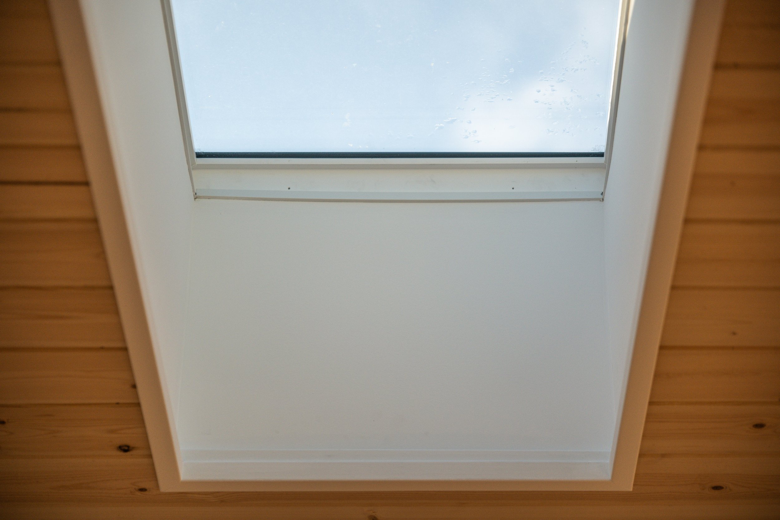Ceiling skylight window with glass and surrounding white frame, some raindrops on the glass, wooden ceiling surrounding the window.