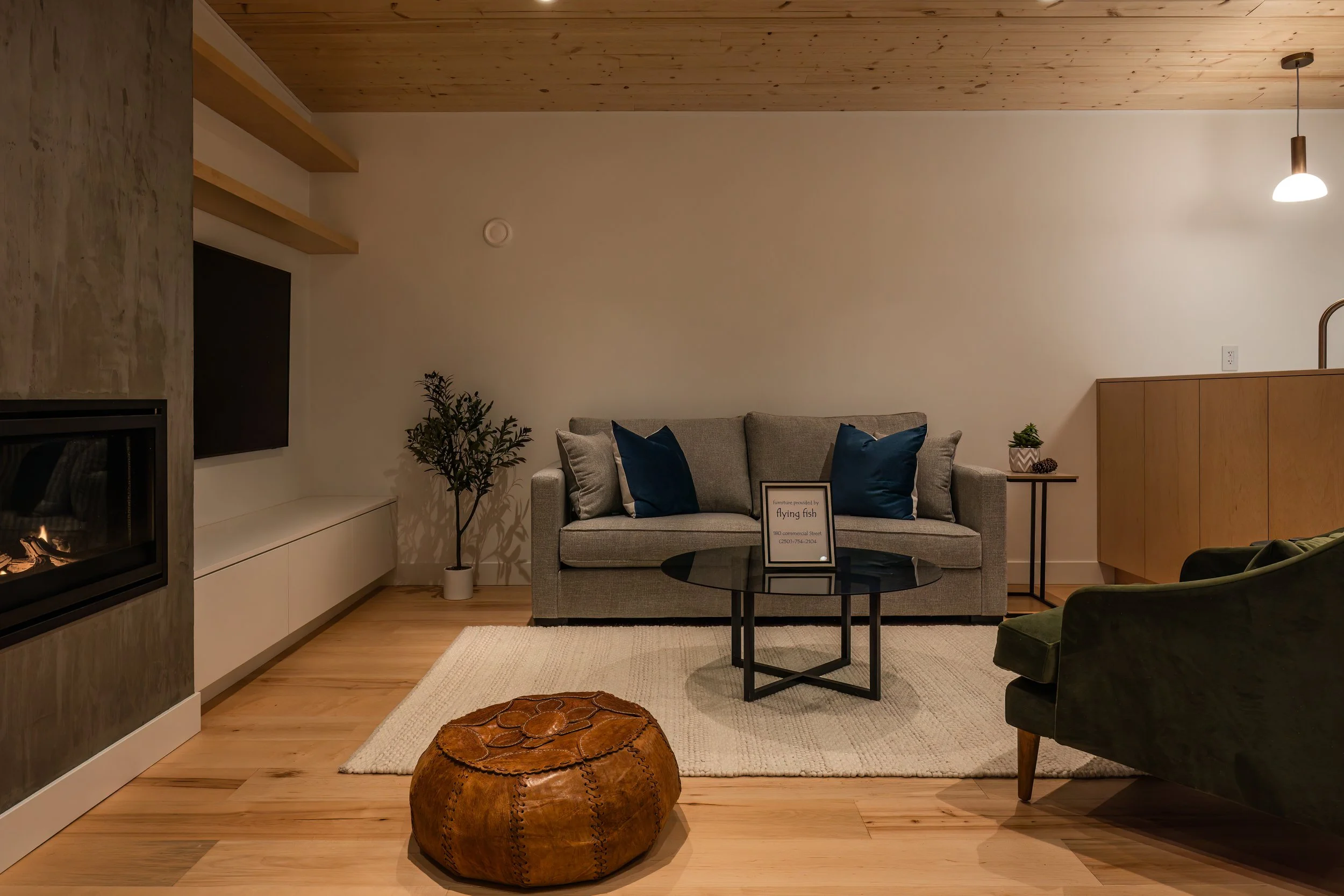 Living room with a gray sofa, navy pillows, a glass coffee table, a potted plant, green armchair, a fireplace, a wooden ceiling, a floor lamp, and a white wall.