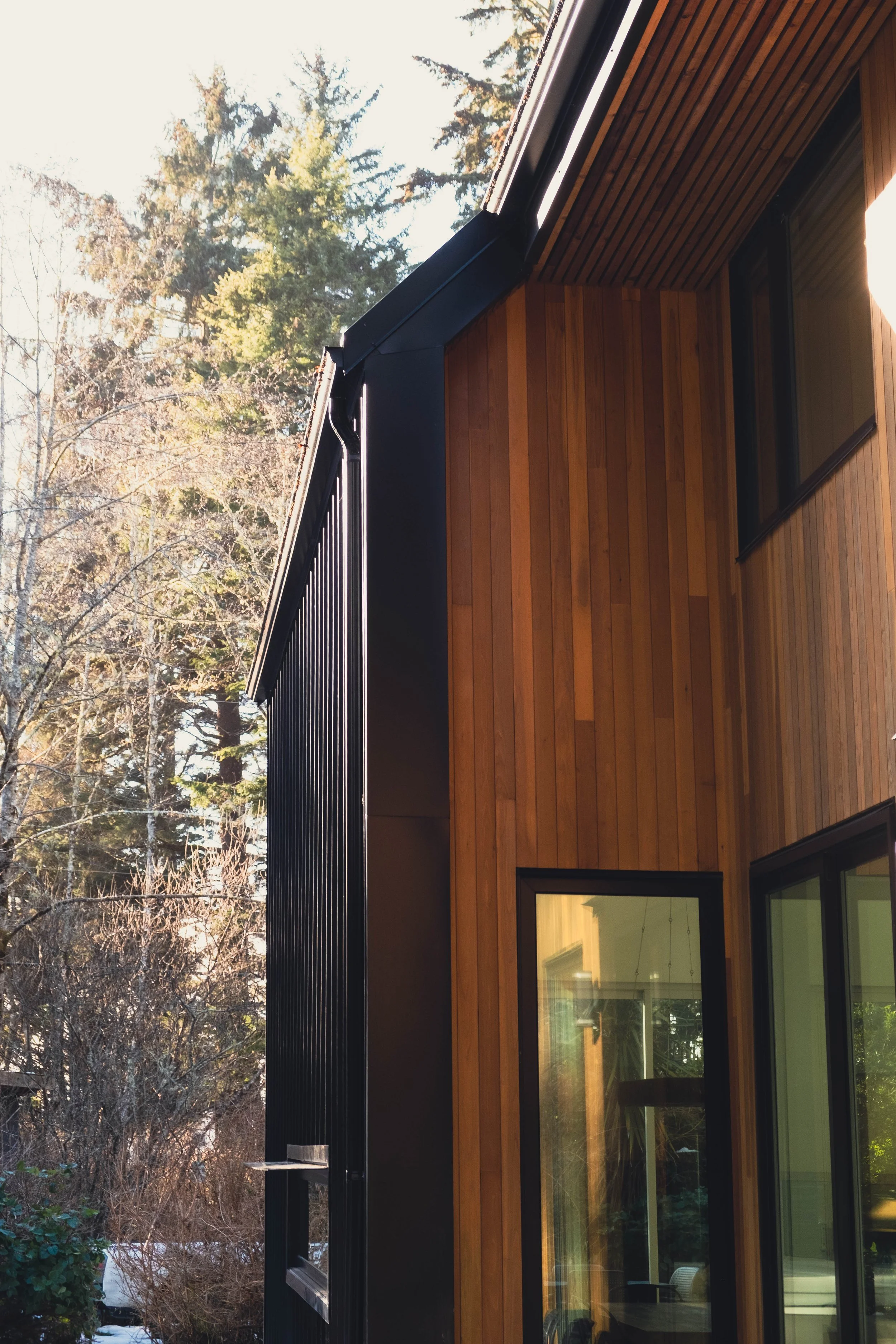 Side view of a modern house with wooden siding and large windows, surrounded by trees.
