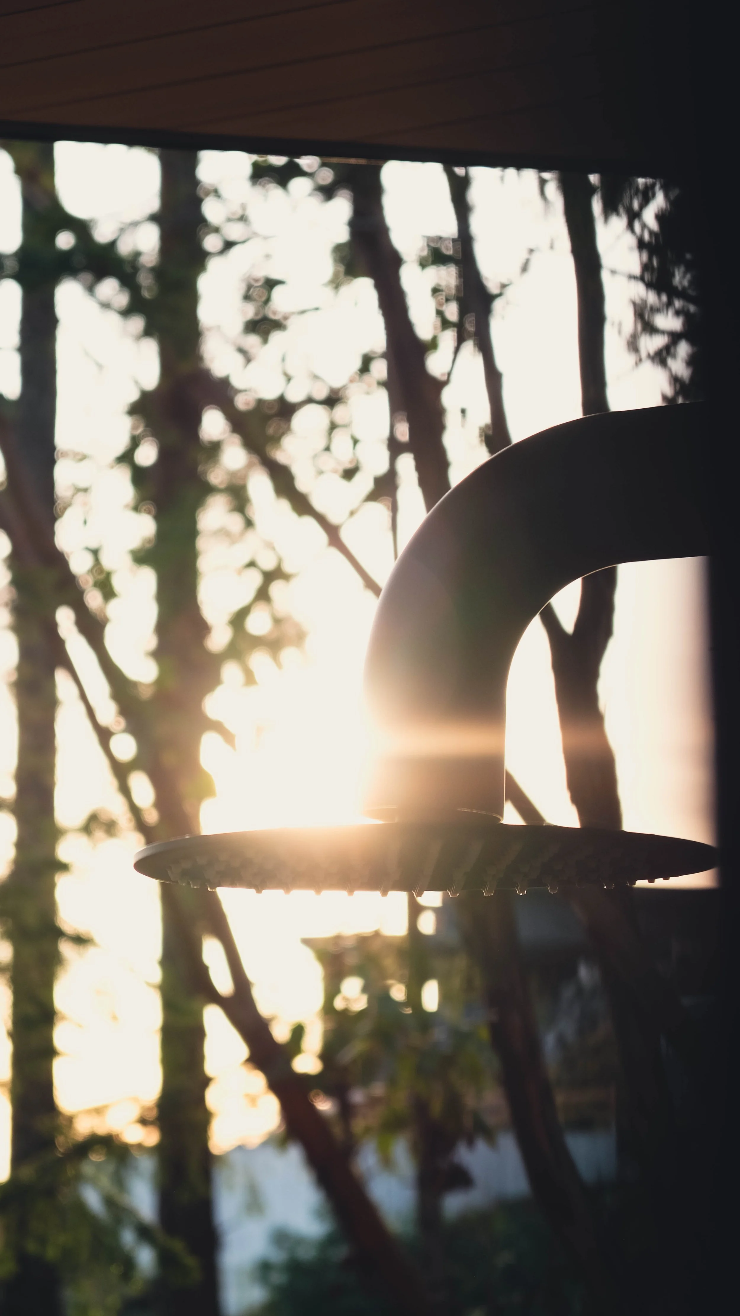Close-up of a showerhead attached to a dark structure with the sunset and trees visible in the background
