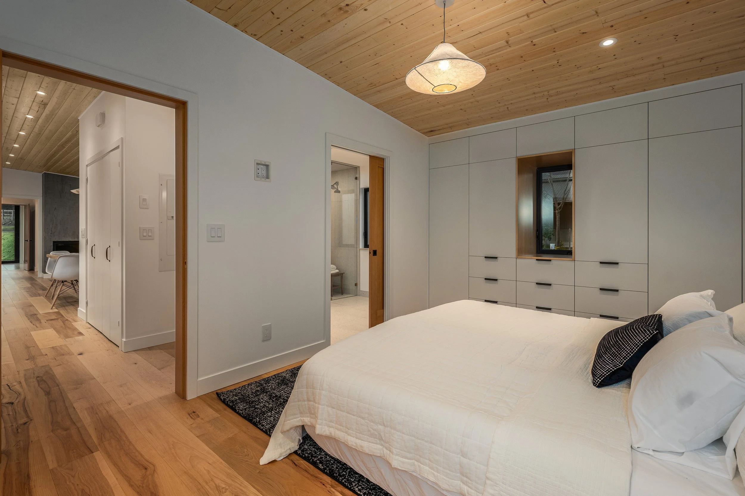 Modern bedroom with a white bed, wooden ceiling, and built-in cabinetry with a window. Adjacent to a bathroom and hallway.