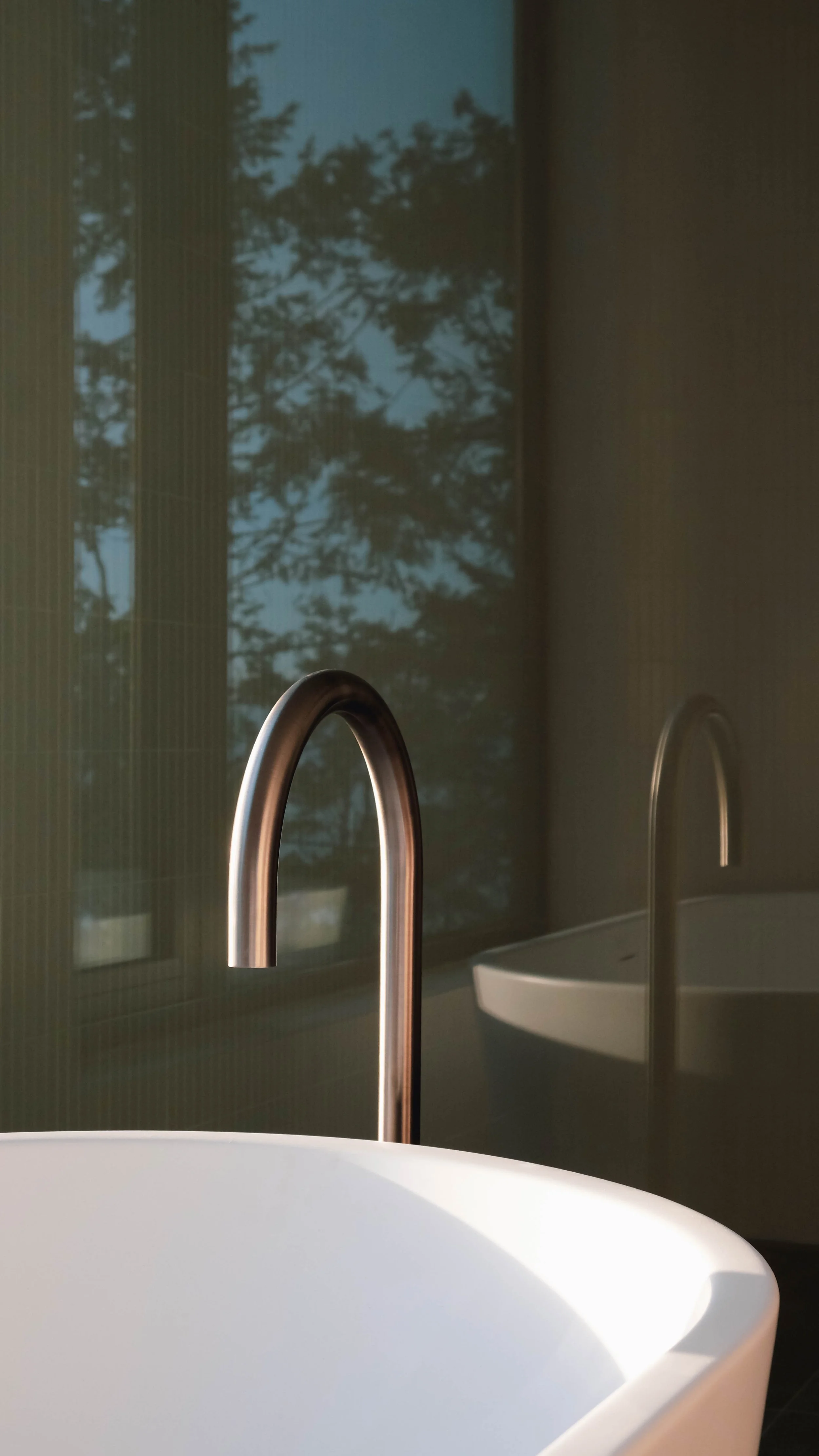 Close-up of a modern sink with a sleek curved faucet, reflection of the faucet can be seen in a mirror, with a large window showing trees outside in the background.