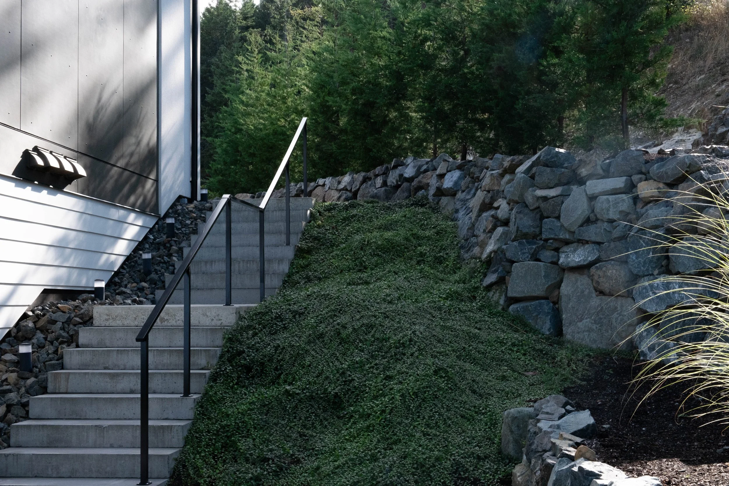 An outdoor concrete staircase with black metal railings beside a house, leading up past green shrubbery and rock retaining wall, with trees in the background.