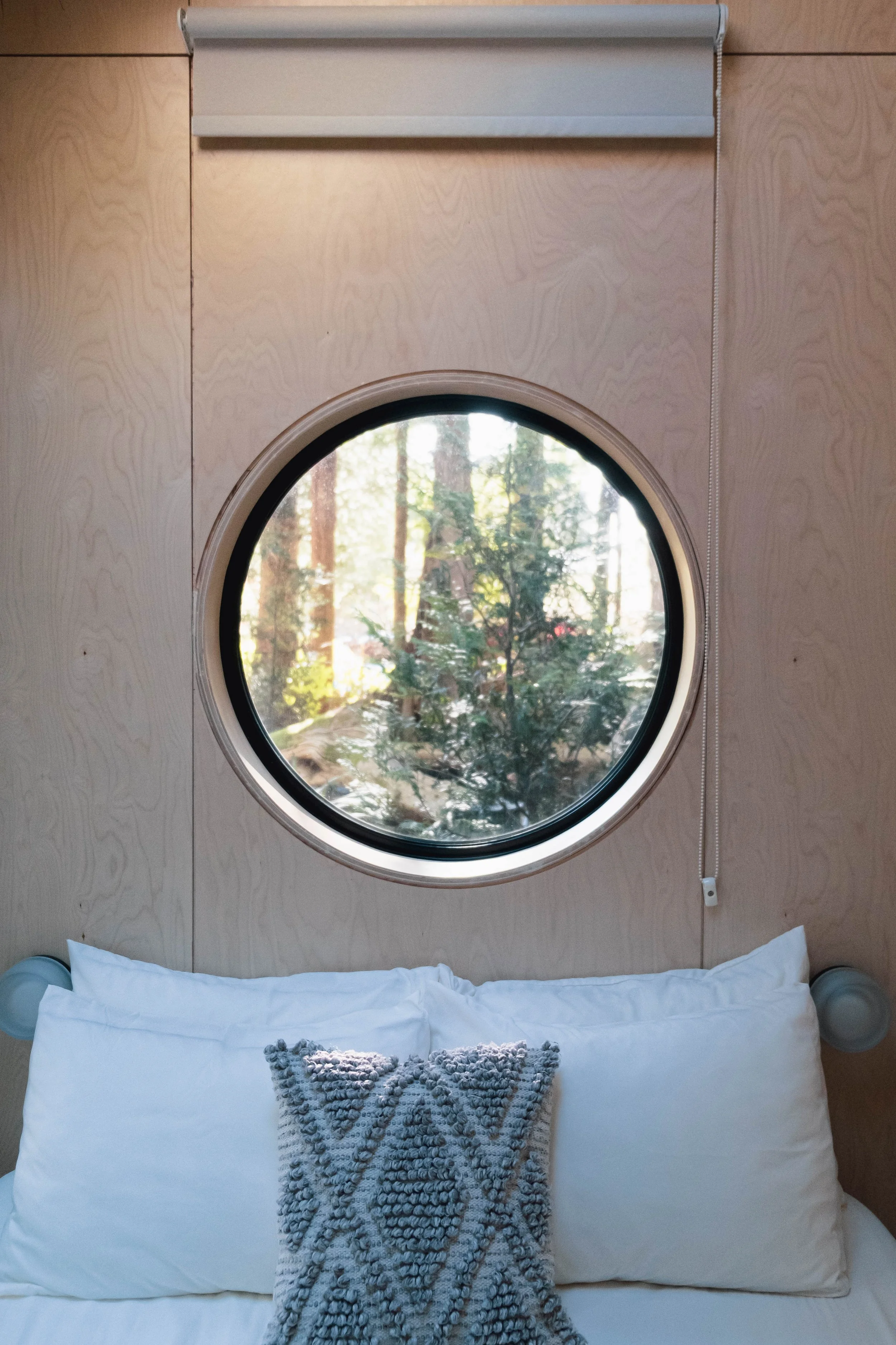 A circular window showing a view of trees outside, above a bed with white pillows and a textured gray throw pillow. The wall is light wood and there is a white roller shade at the top.