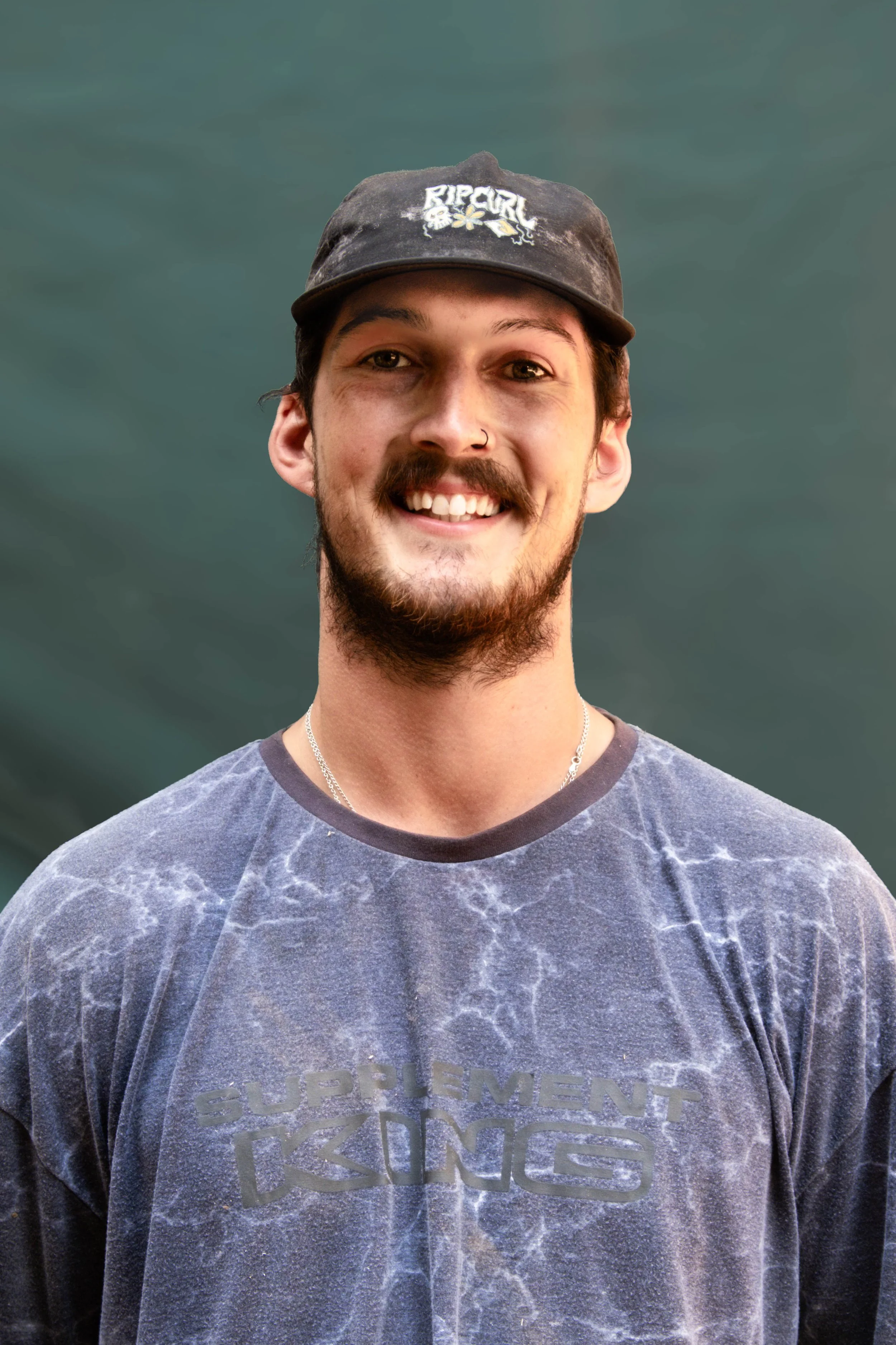 A young man with a beard and mustache smiling, wearing a black cap and a blue t-shirt, standing outdoors with a blurred green background.