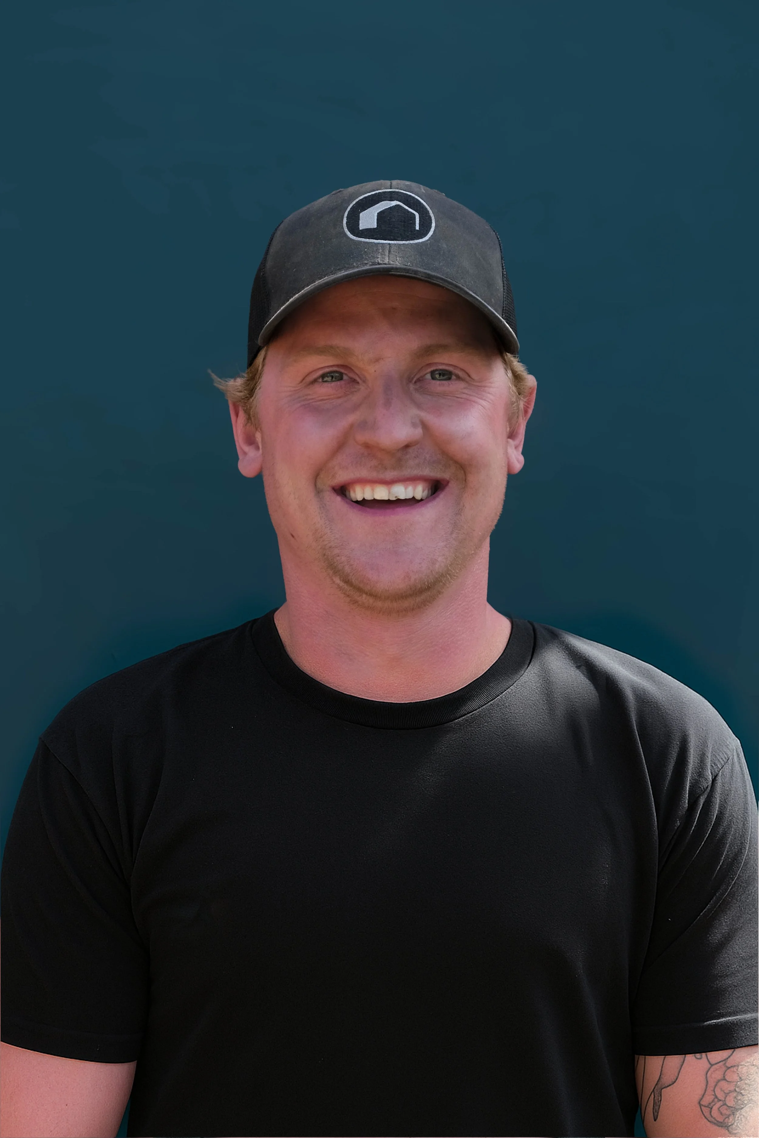 A smiling man wearing a black baseball cap and black t-shirt against a dark blue background.