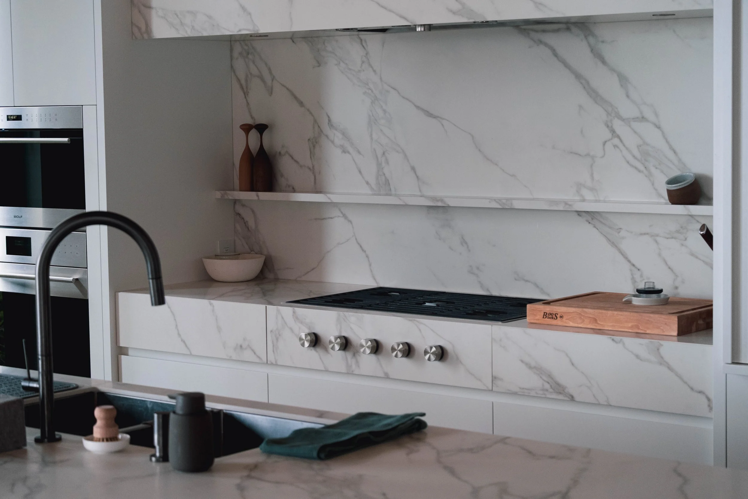 Modern kitchen with white marble countertops and backsplash, stainless steel oven, stovetop, and shelves with decorative items.