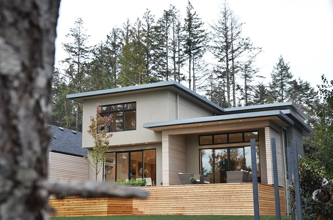 Modern two-story house with large glass windows and sliding doors, wood deck, and surrounded by tall trees.