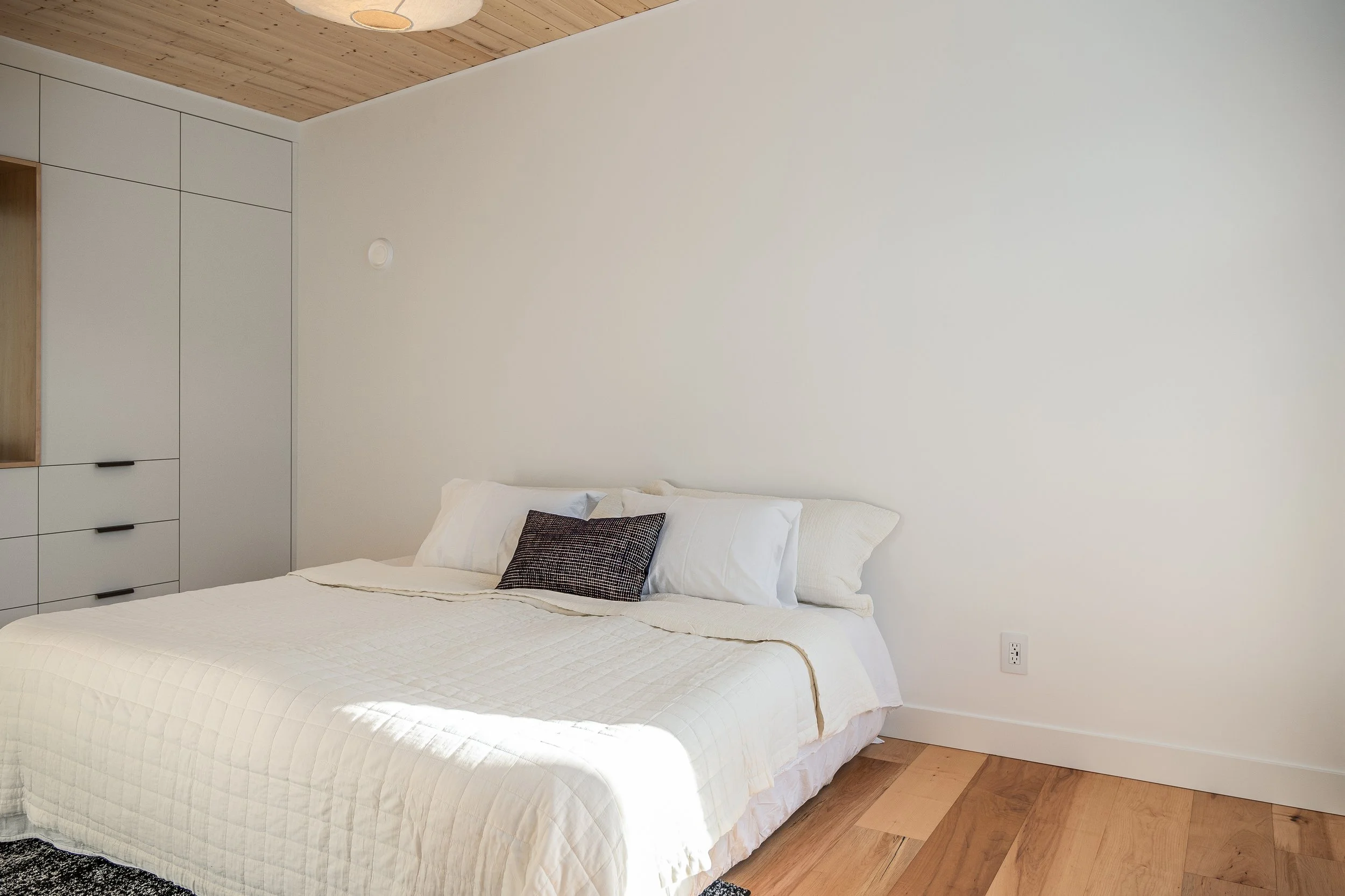 A neatly made bed with white bedding and pillows, featuring a dark patterned pillow, in a minimalist bedroom with white walls, wooden flooring, and a built-in wardrobe.